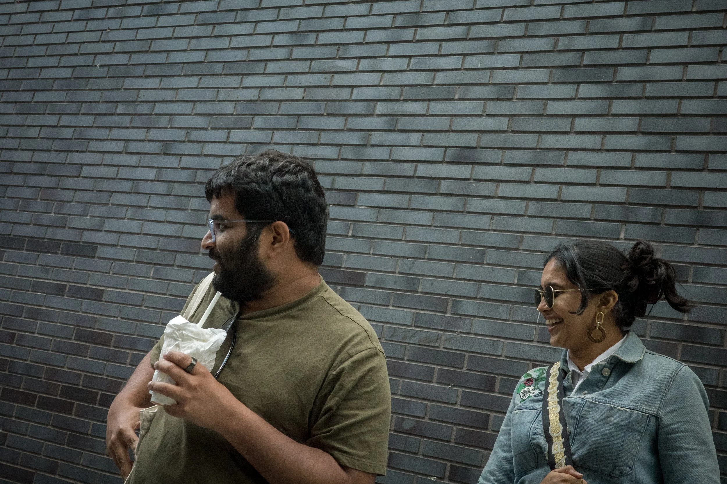 Two people laughing and chatting against a black brick wall, one holding a crumpled paper bag, the other wearing sunglasses and earrings.