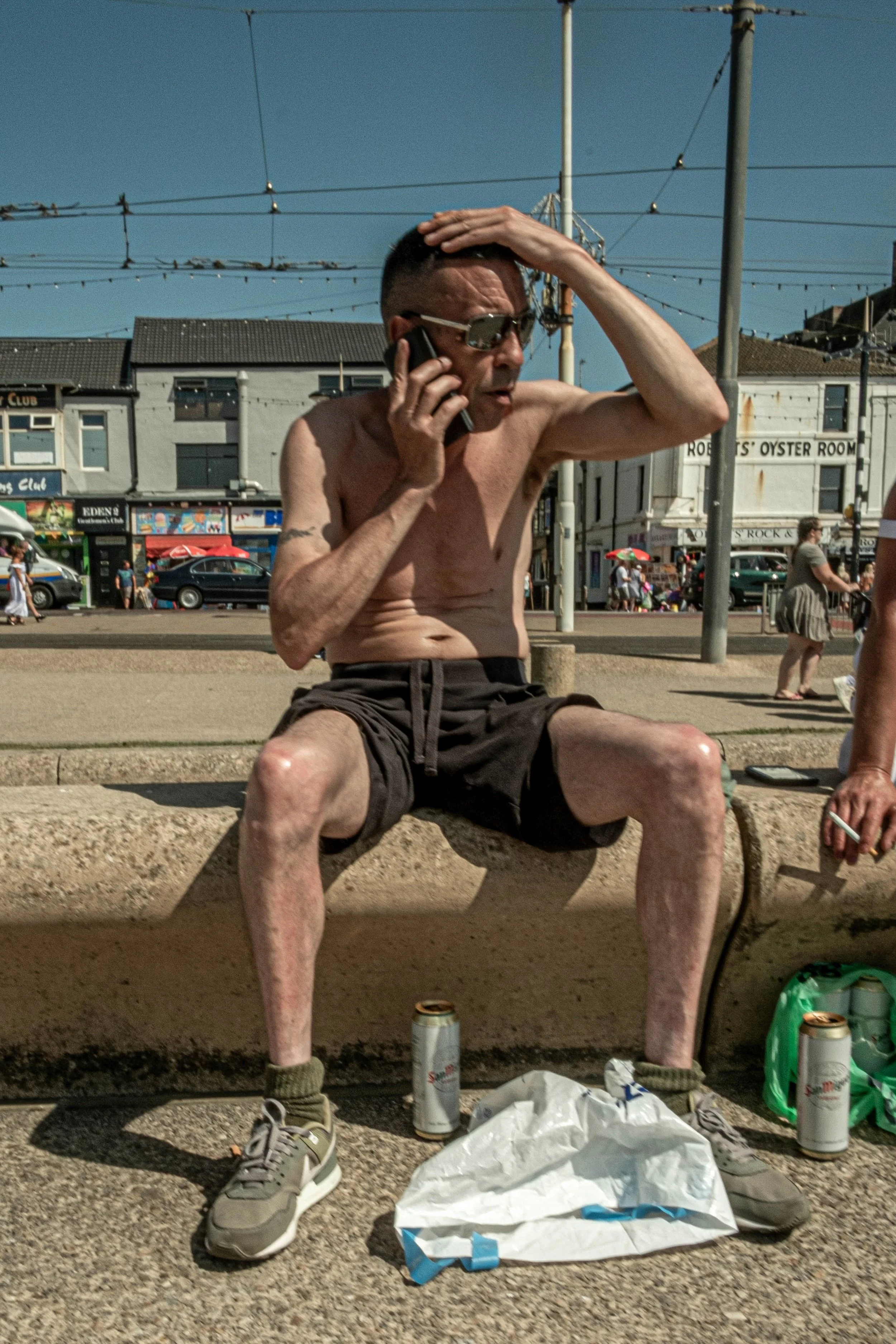 A shirtless man wearing sunglasses is sitting on a bench at a beach, holding a phone to his ear, with one hand on his head. Surrounding him are various items including two cans, a green bag, sneakers, and a white plastic bag. In the background, peopl