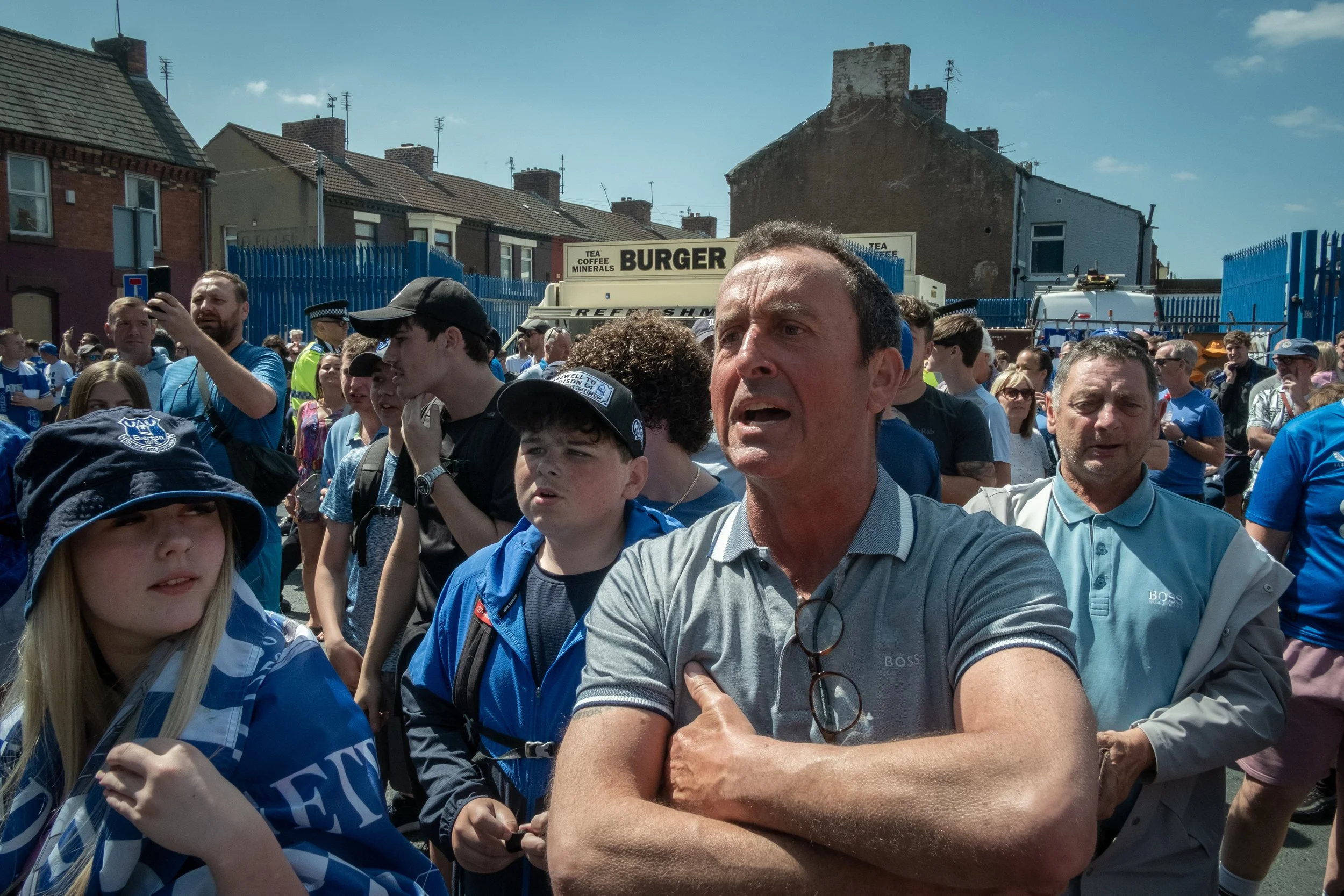 Crowd of people gathered outdoors, some wearing blue clothing, with a man in the foreground looking emotional and others taking photos or watching.