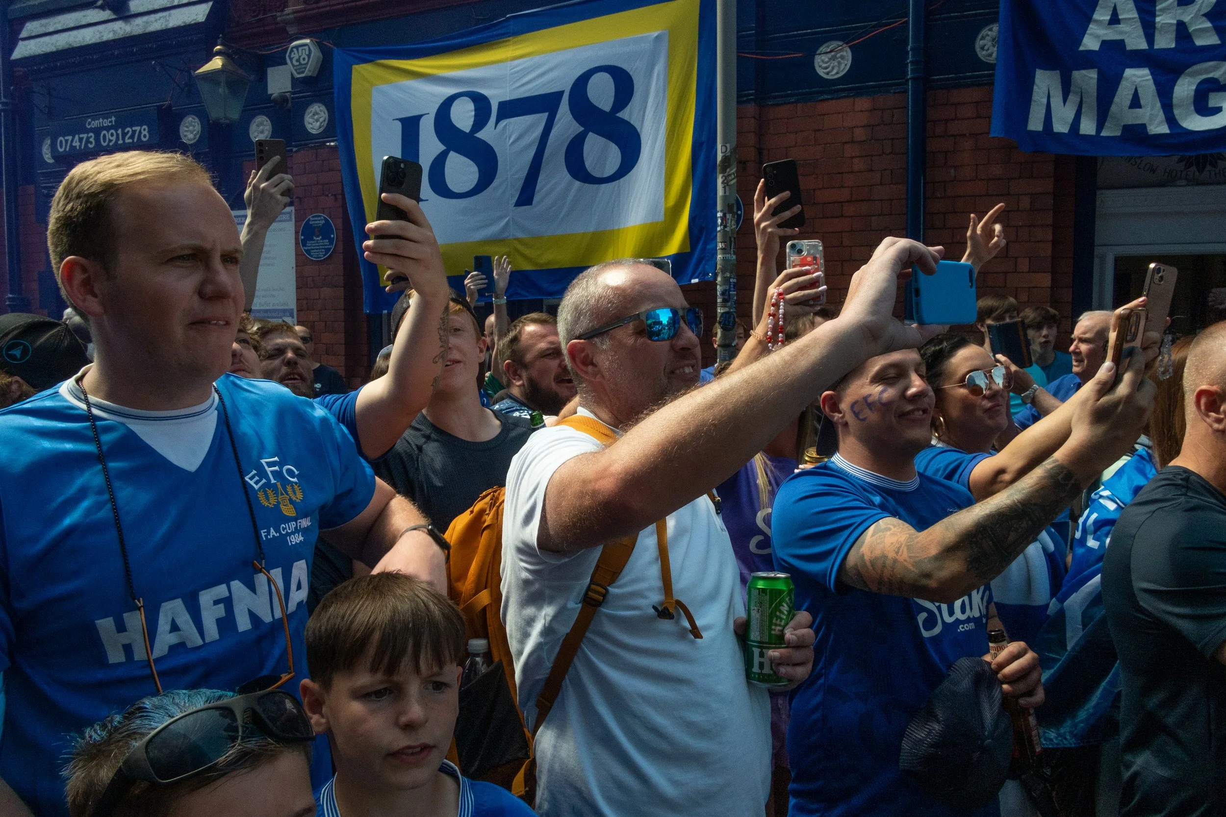A group of football fans gathering outdoors, some wearing blue team jerseys, taking photos with smartphones, with a large banner reading '1878' in the background.