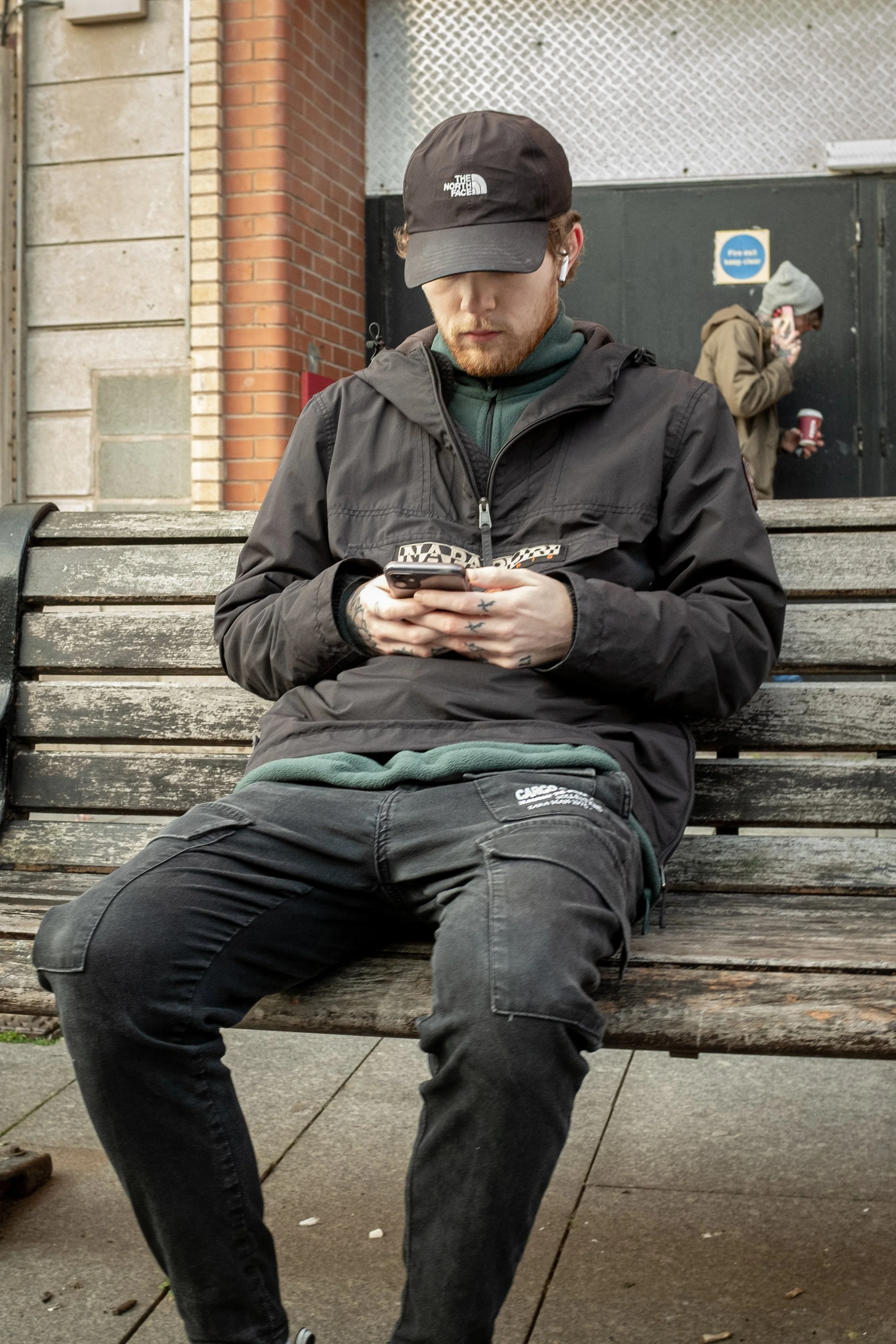 A young man with a red beard sitting on a wooden park bench, wearing a black cap, black jacket, and dark pants, looking at his phone. In the background, a person in a gray hoodie is smoking and holding a red cup.