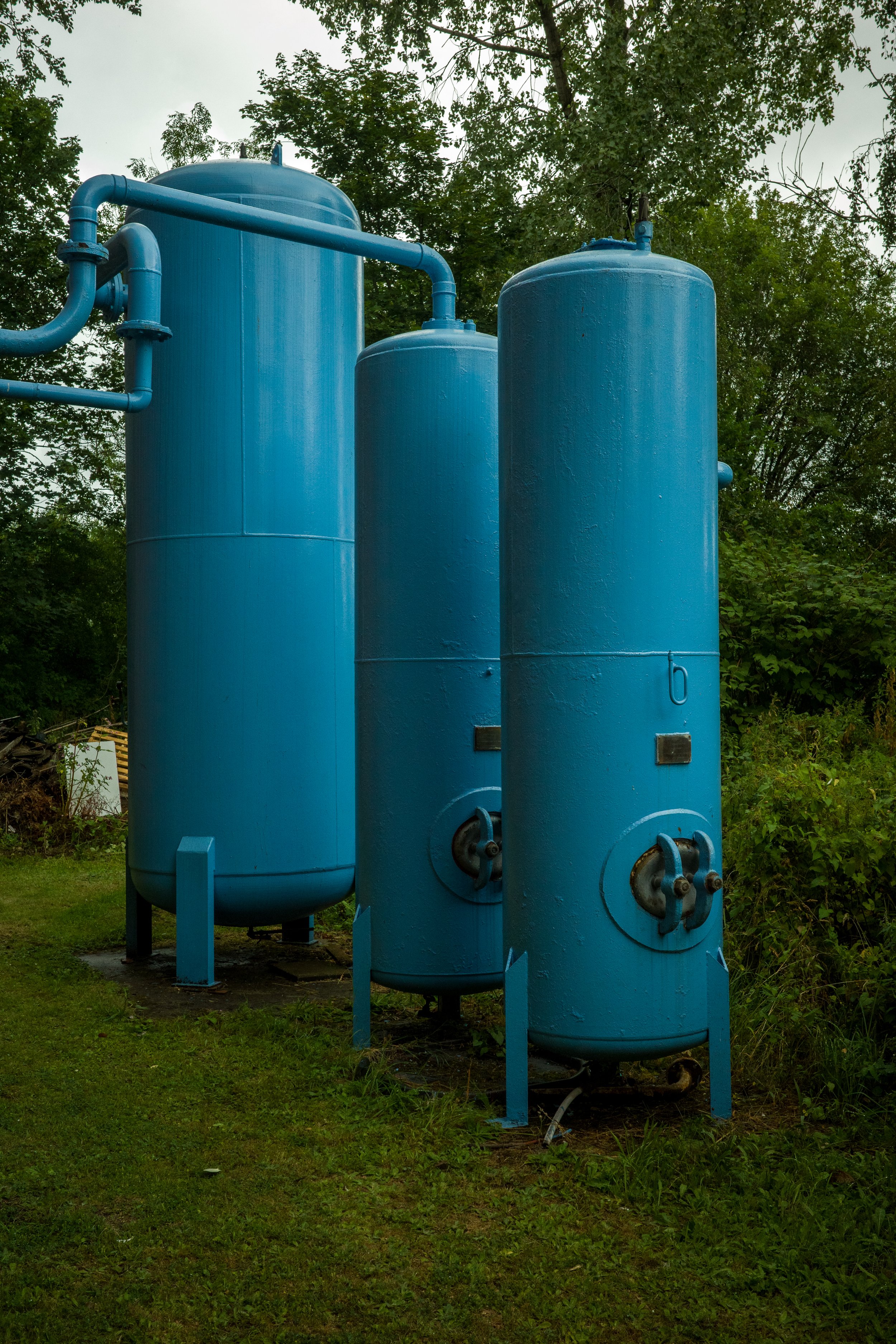 Three large, blue industrial tanks with pipes on top, situated outdoors on a grassy area with trees in the background.