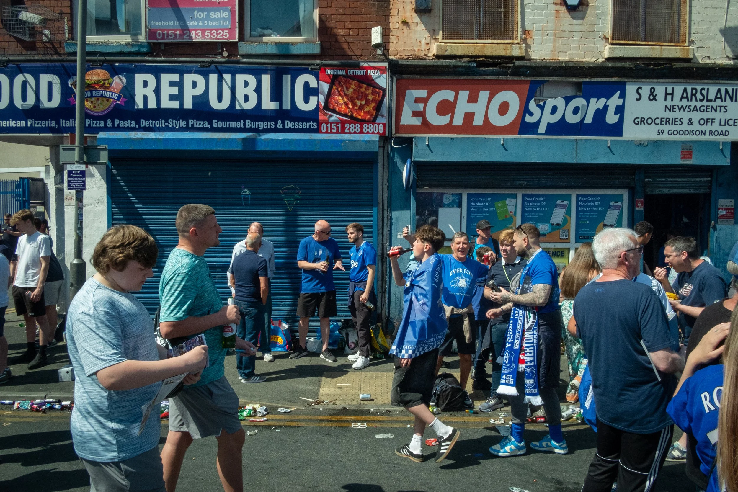 People gathering on a street, many wearing blue sports jerseys representing Everton Football Club, outside storefronts with signs in the background.