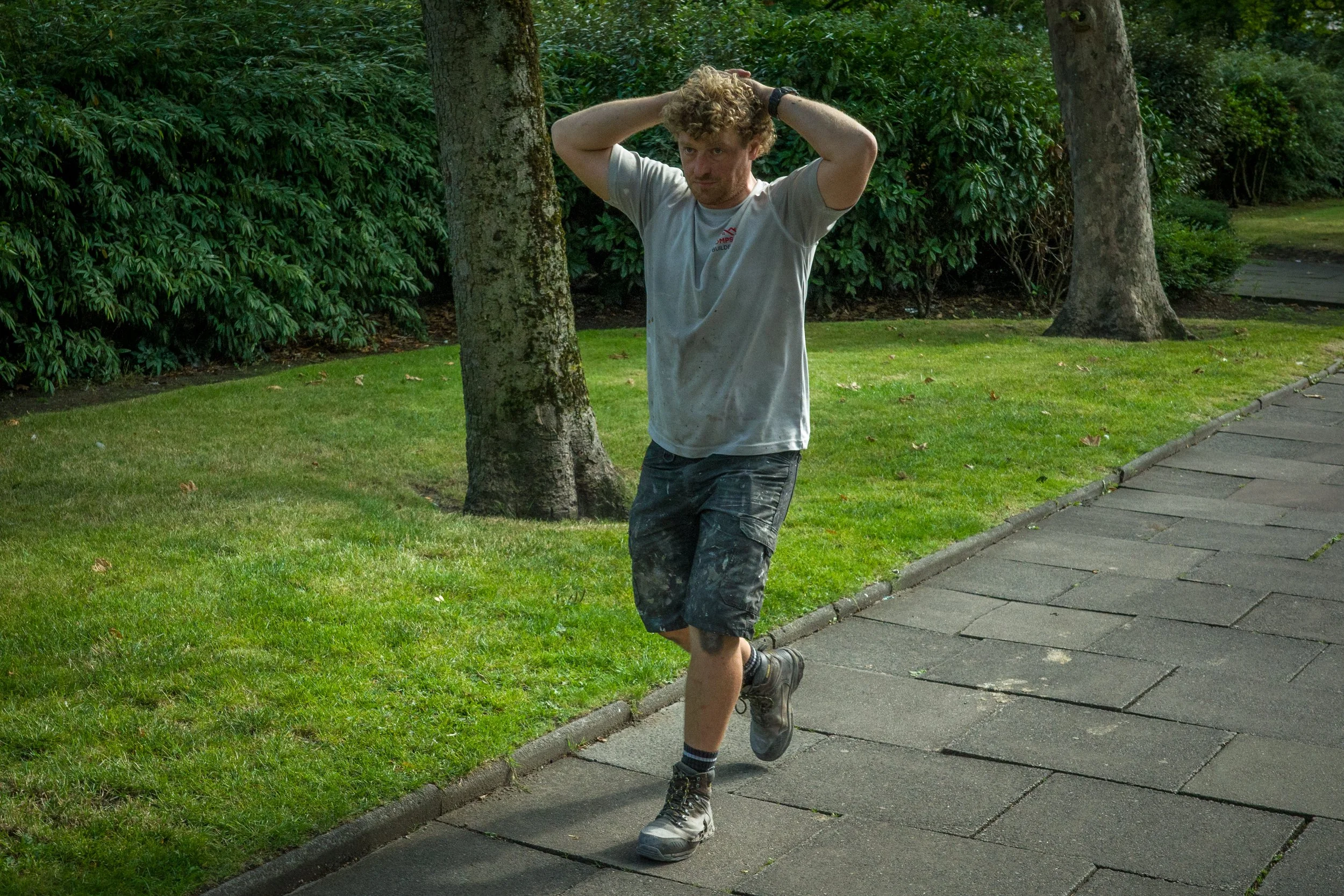 A man with curly hair and a beard walking outdoors on a concrete sidewalk next to a grassy area and trees, wearing a dirty white t-shirt, black shorts, and boots, with his hands behind his head.