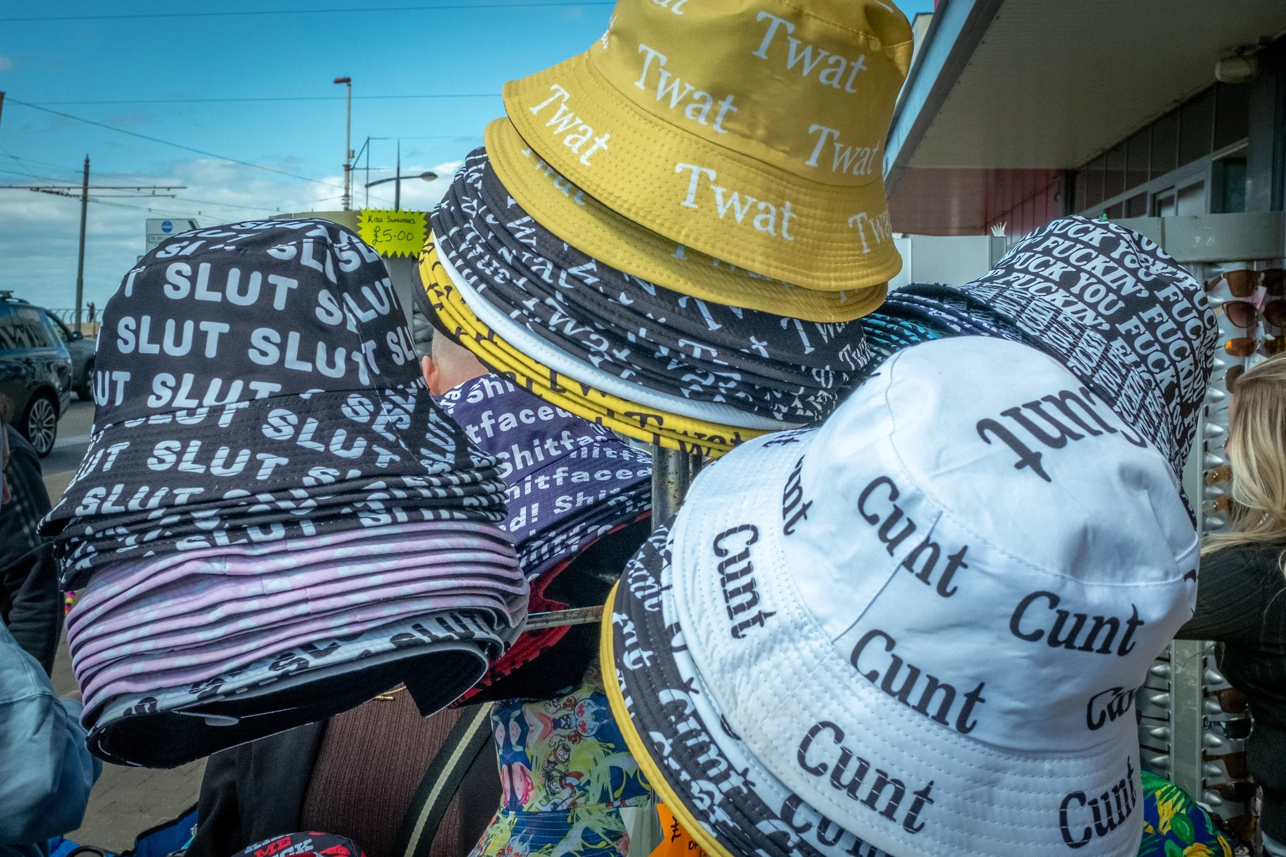 A display of hats with offensive and explicit words in various colors and styles at an outdoor market. Some hats are printed with words like 'SLUT,' 'Cunt,' and 'FUCK,' while others have the word 'Twat.'