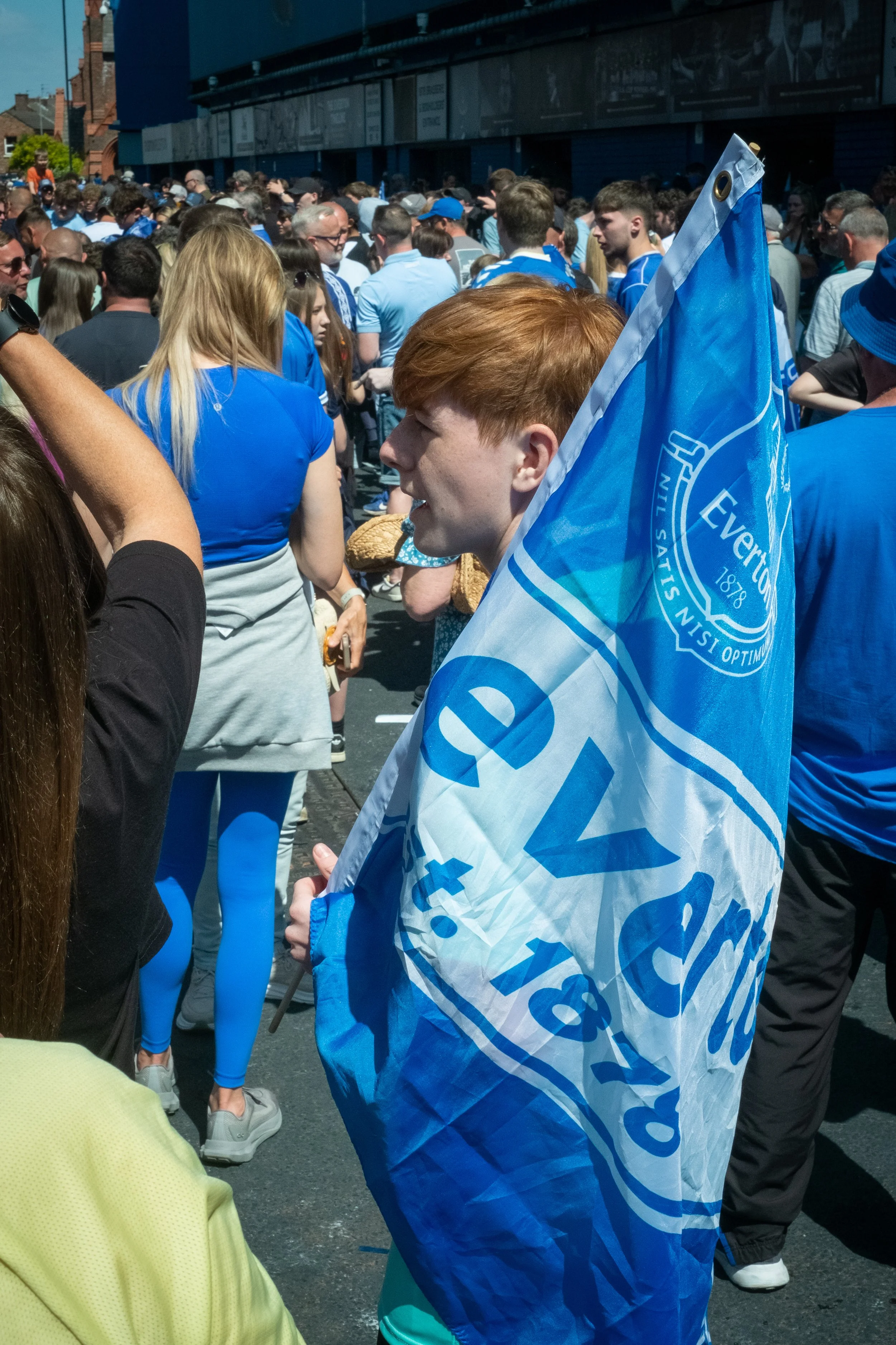 A crowd of people gathered outdoors, with someone holding an Everton F.C. flag. The crowd includes individuals wearing blue, some in athletic attire, in front of a building with large windows.