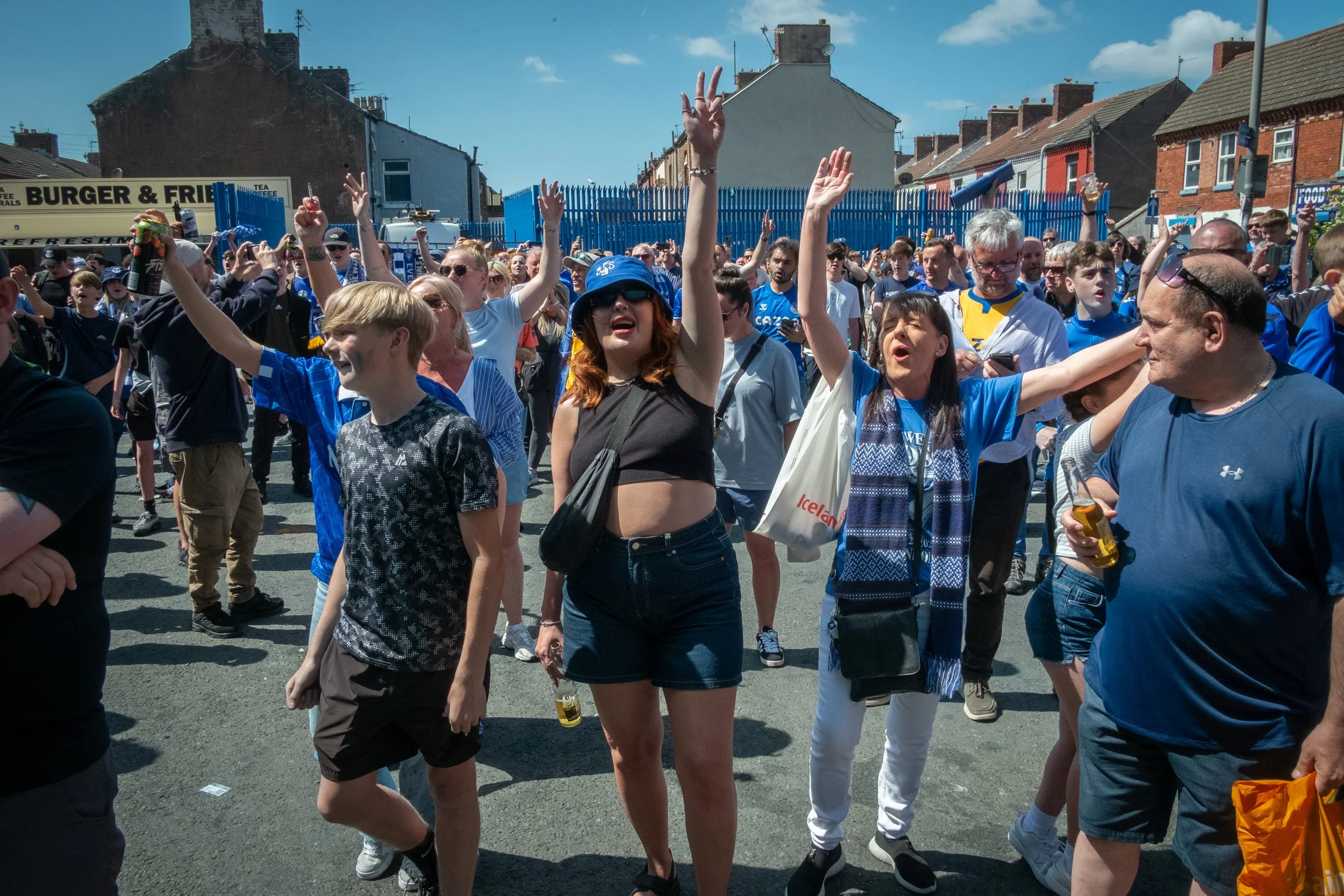 Crowd of sports fans outdoors, many wearing blue, cheering with hands raised, with blue fence and houses in background on sunny day.