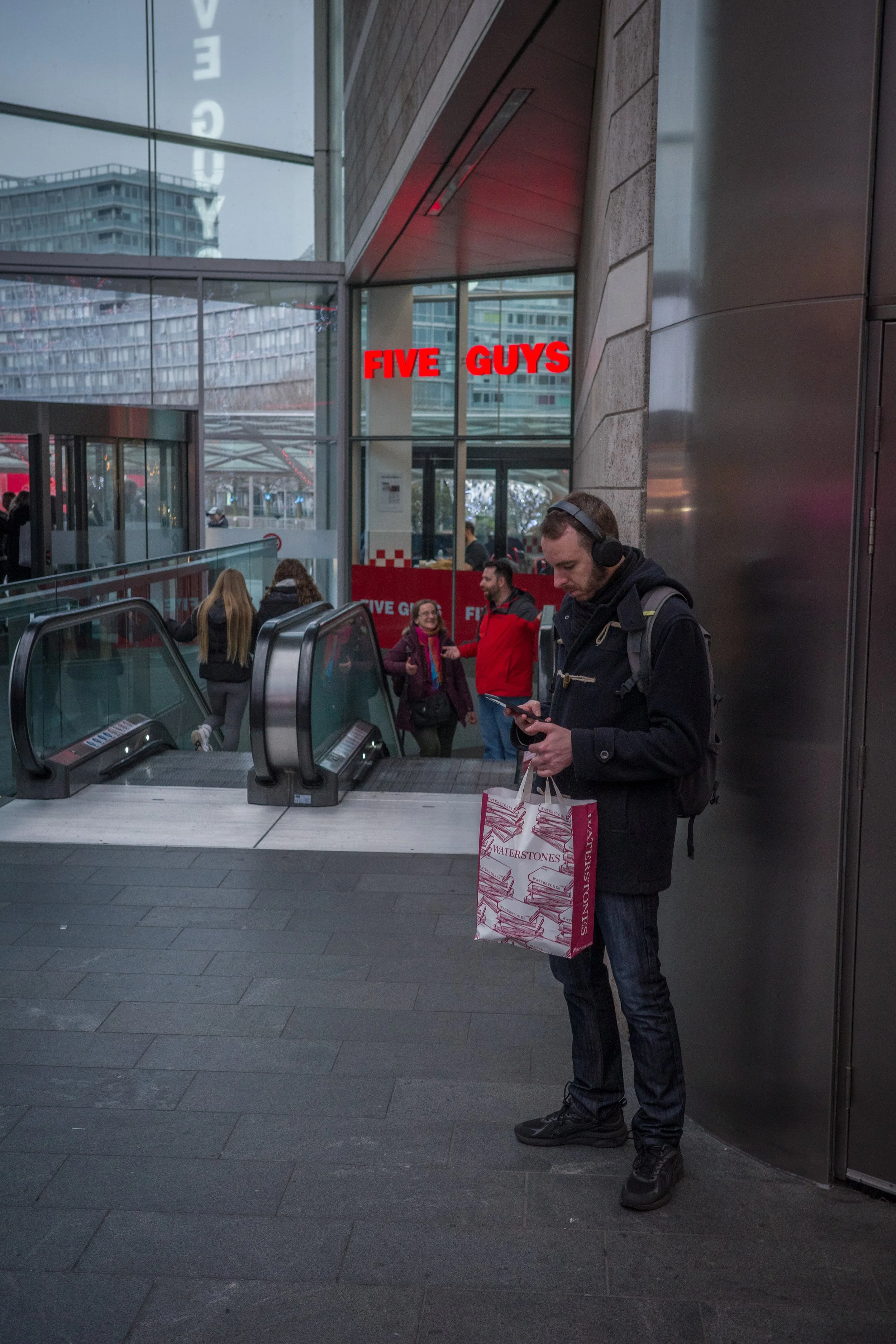 People waiting at an escalator inside a modern building with glass walls. A man in the foreground is looking at his phone while holding a paper shopping bag, and there are others talking and using their phones in the background. Red neon sign reading