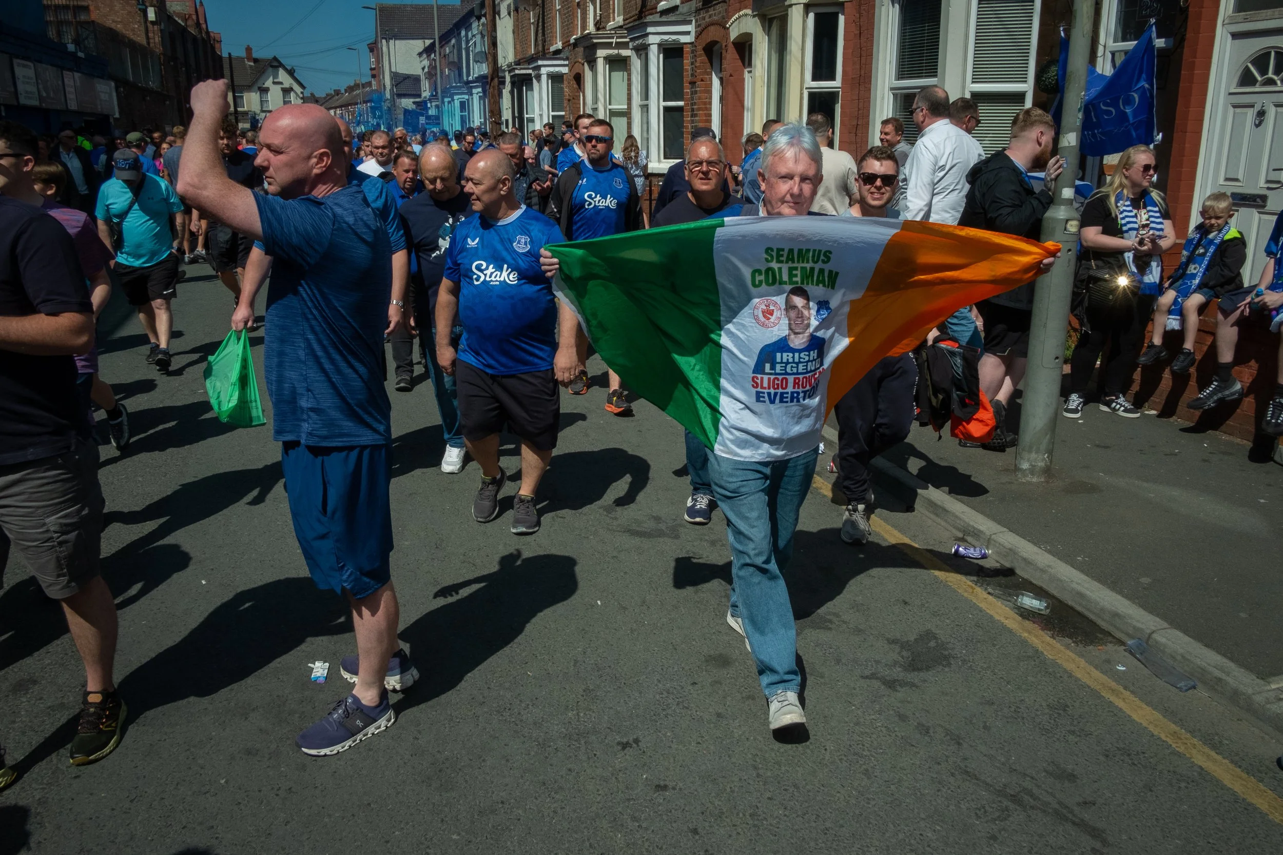 Crowd of people walking down a city street during a parade or protest, with one person holding a flag and wearing a jersey supporting Seamus Coleman for Irish League and Sligo Rovers, on a sunny day.