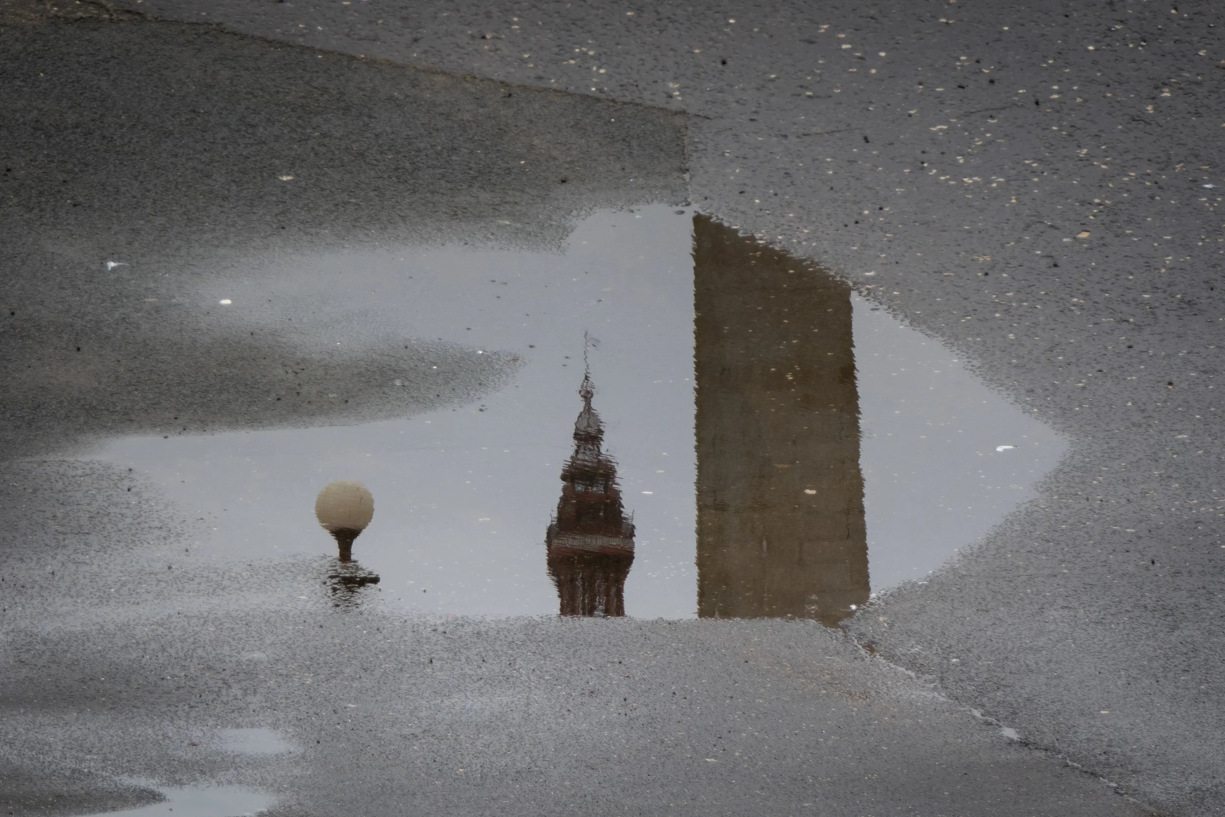 Reflection of a tower and urban streetlight in a puddle on asphalt pavement.