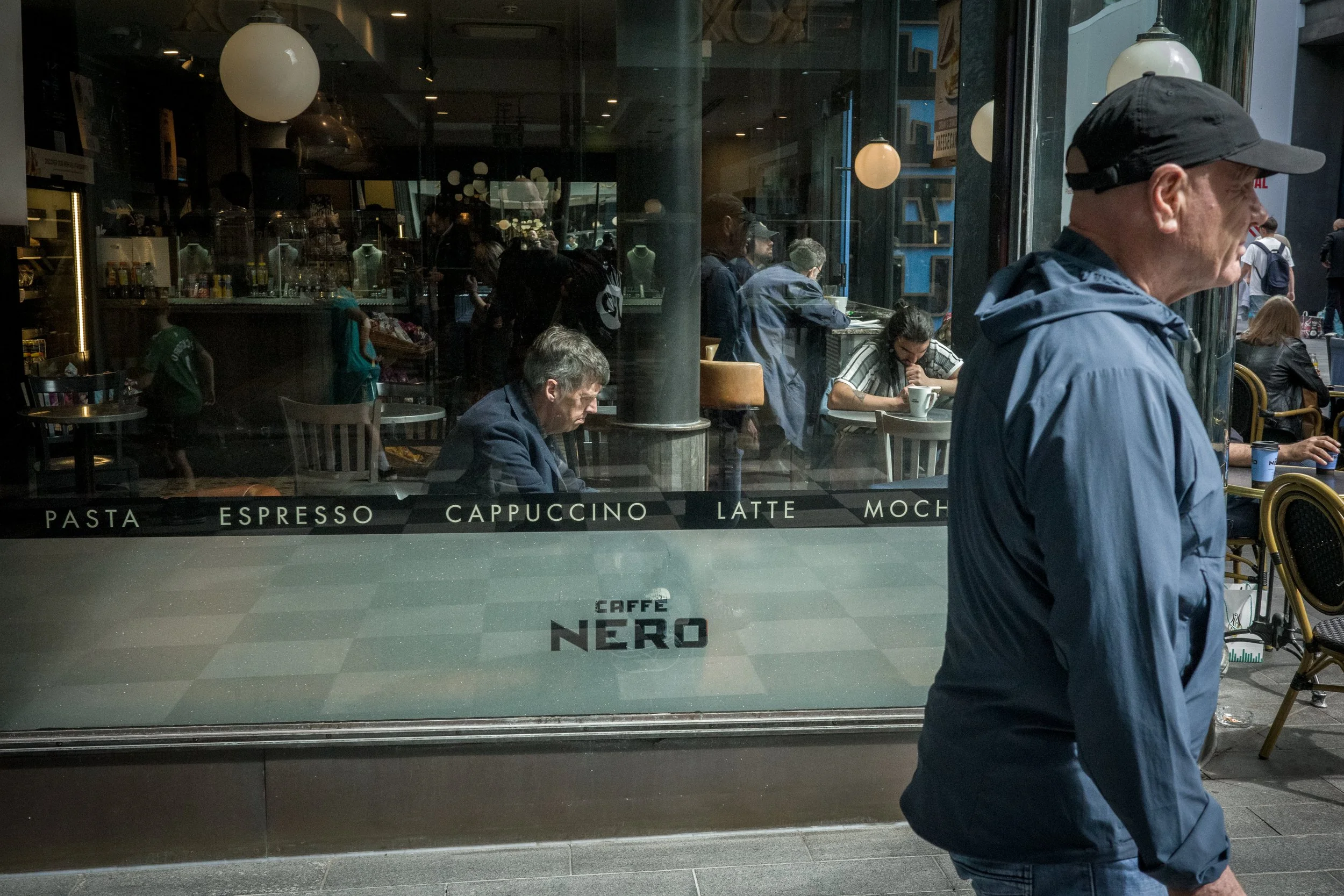 People dining inside a coffee shop, seen through a large window, with one person walking past outside.