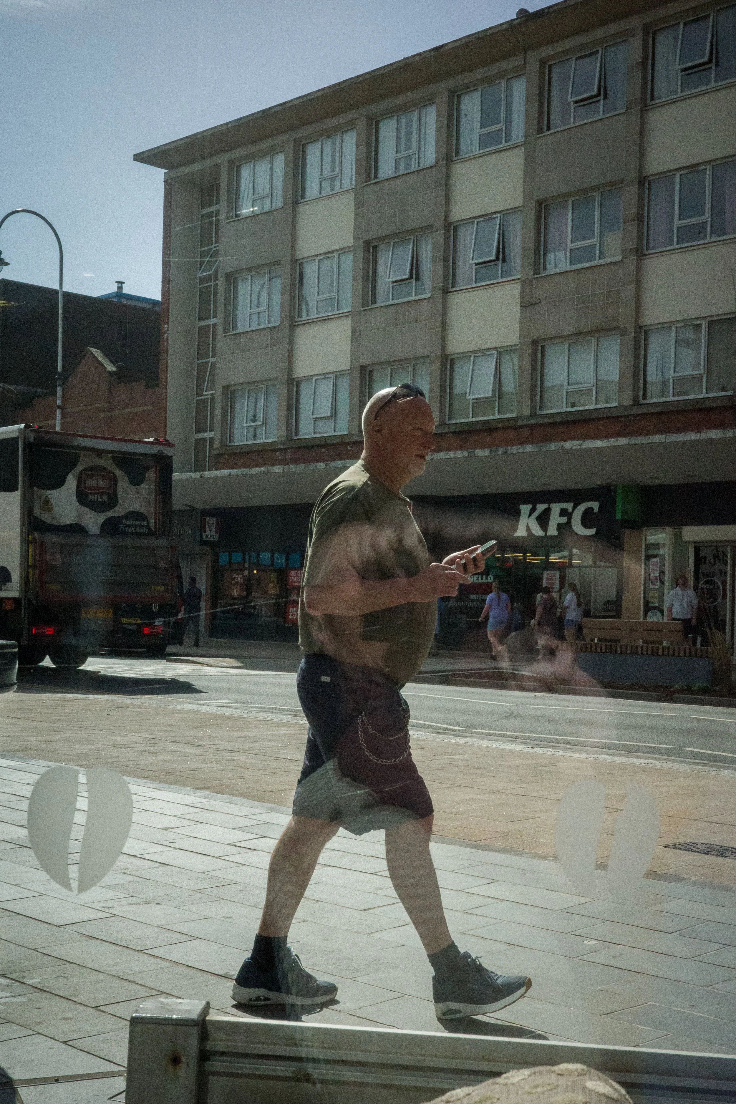 A man in a tan shirt, black shorts, and black sneakers is walking on a city sidewalk while looking at his phone. He is wearing sunglasses on his head. In the background, there is a KFC restaurant and a multi-story apartment building. The photo appear