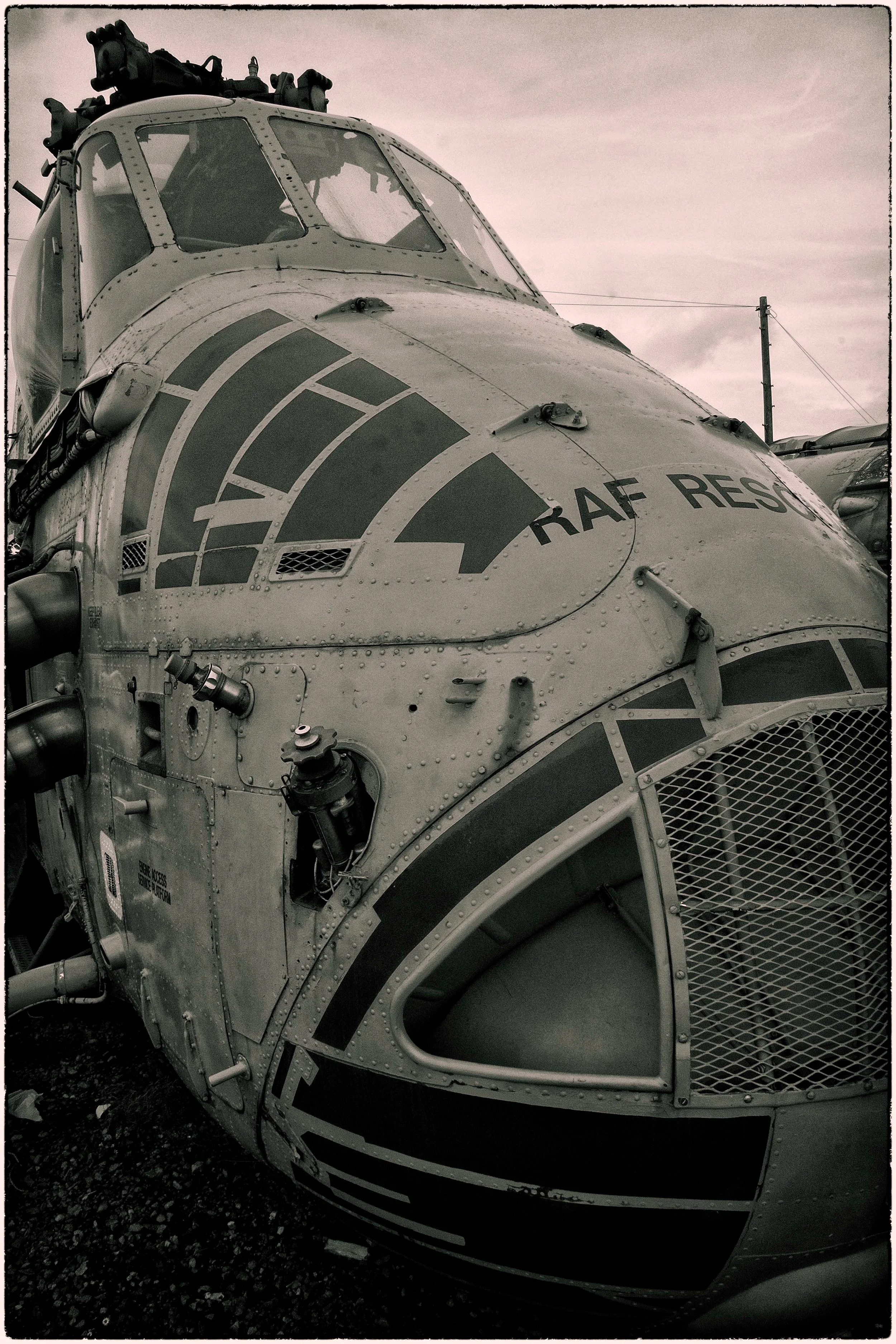 Close-up of a military helicopter with several warning labels, antennas, and equipment on its body, parked outdoors with power lines in the background. The helicopter has a rugged, metallic appearance.