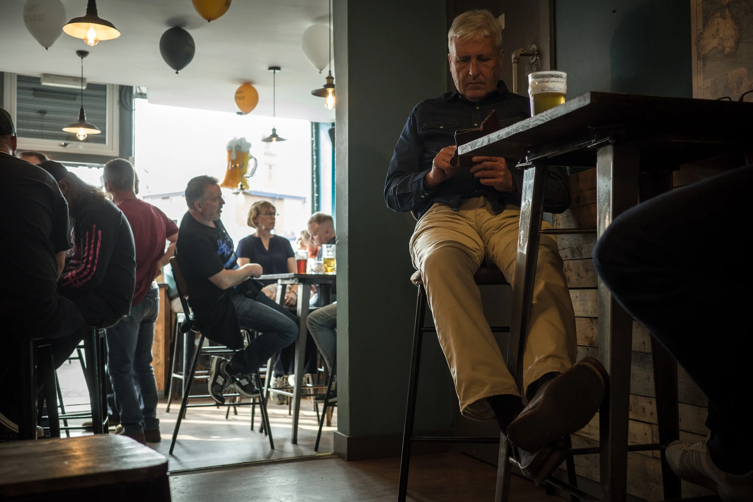 A man with blond hair wearing a dark shirt and beige pants is sitting on a high stool at a bar or restaurant, looking at his phone, with a tall glass of beer in front of him. There are several other people in the background, some sitting and some sta