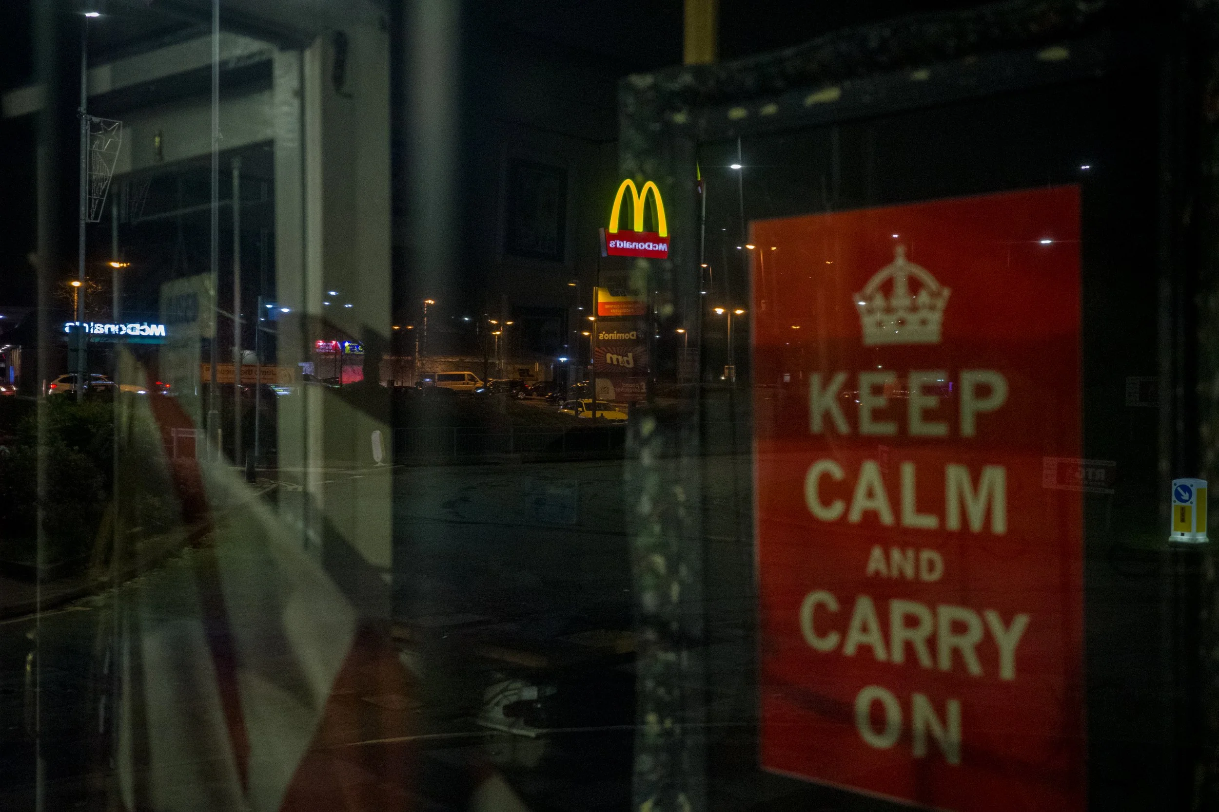 Nighttime view of a McDonald's sign with the golden arches and McDonald's branding visible through a window. There is also a red sign that says "Keep Calm and Carry On" partially visible.