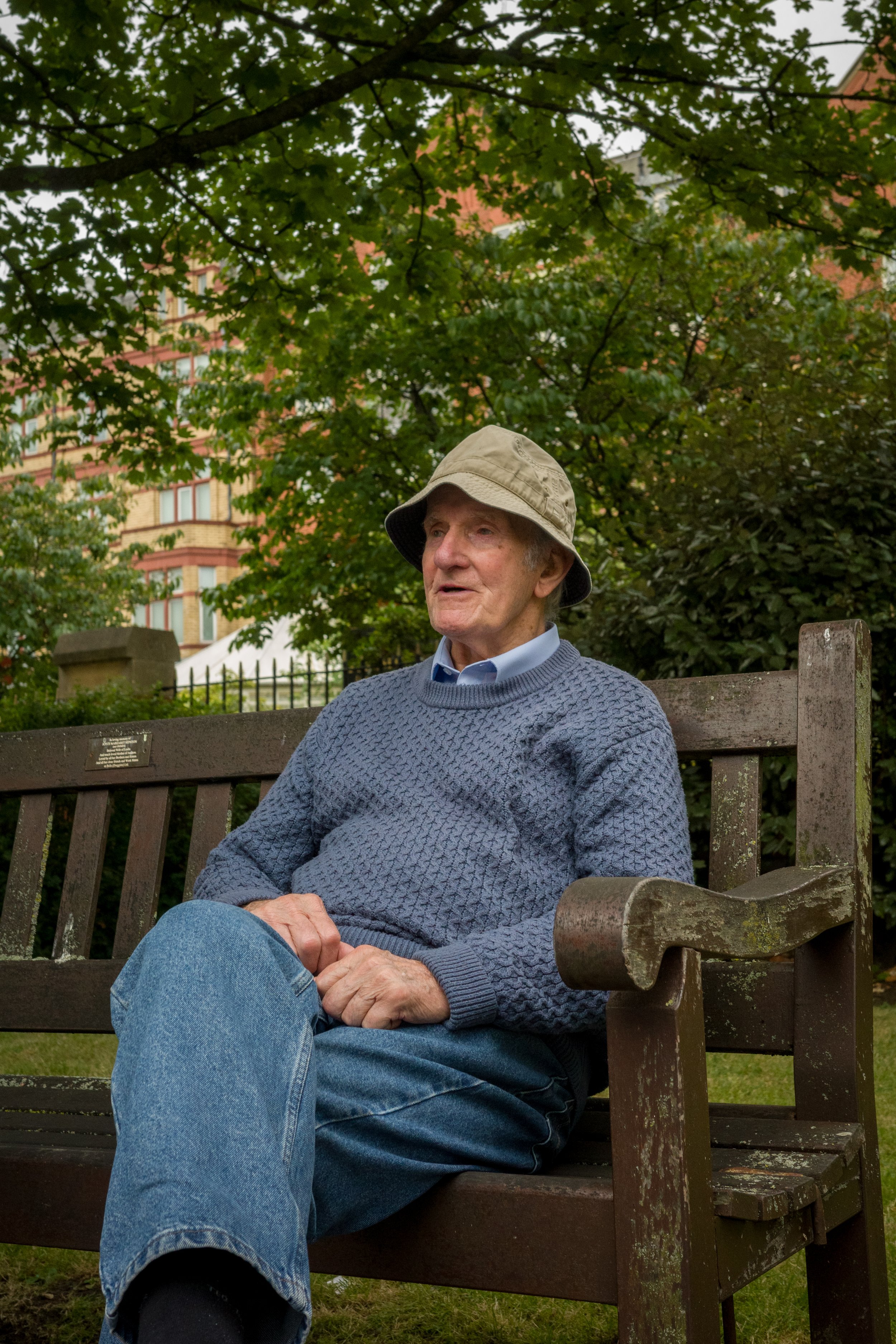 An elderly man sitting on a park bench with trees and a building in the background.