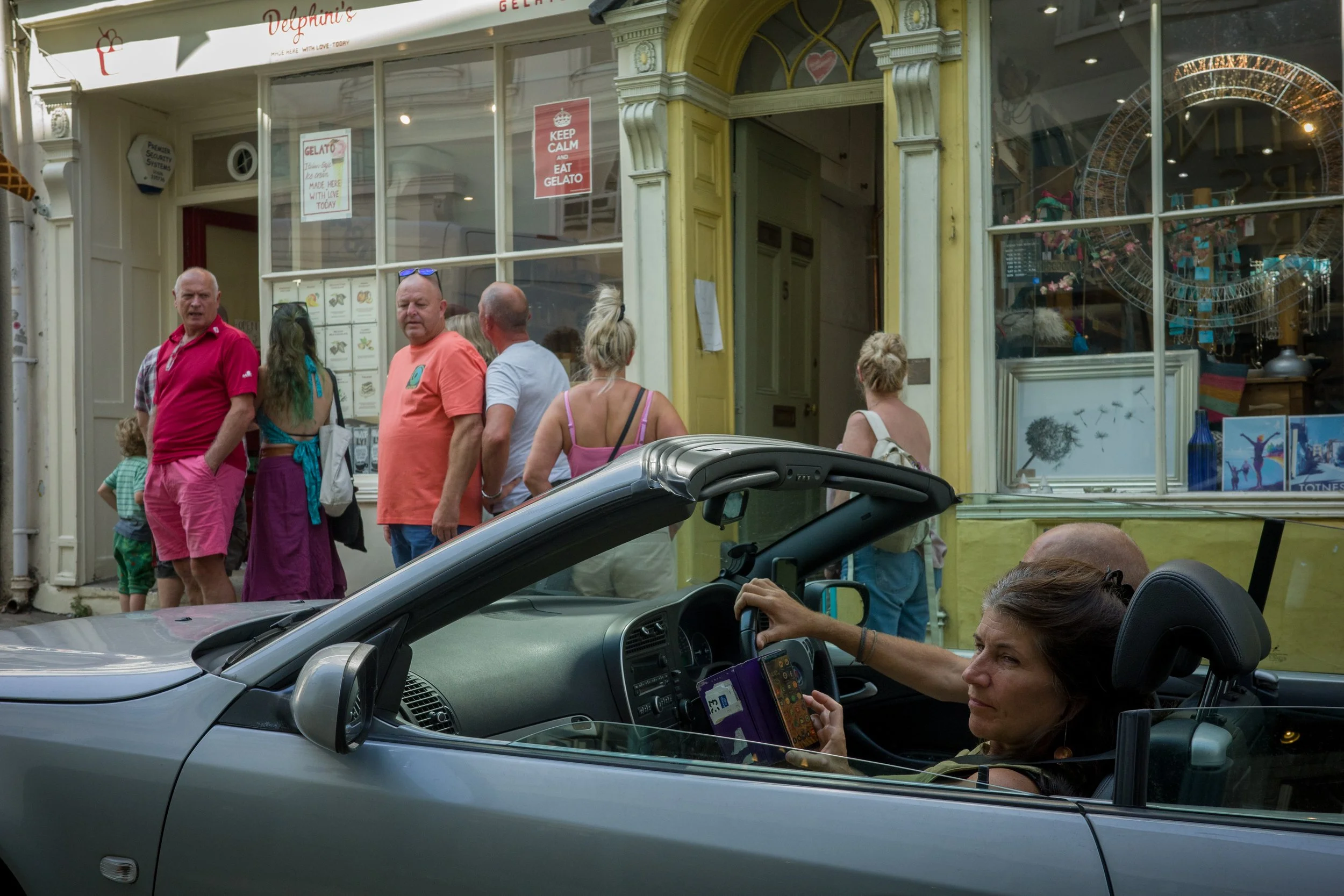 People standing outside a gelato shop, some chatting and others looking inside, while a woman in a car looks at her phone, in an urban setting.