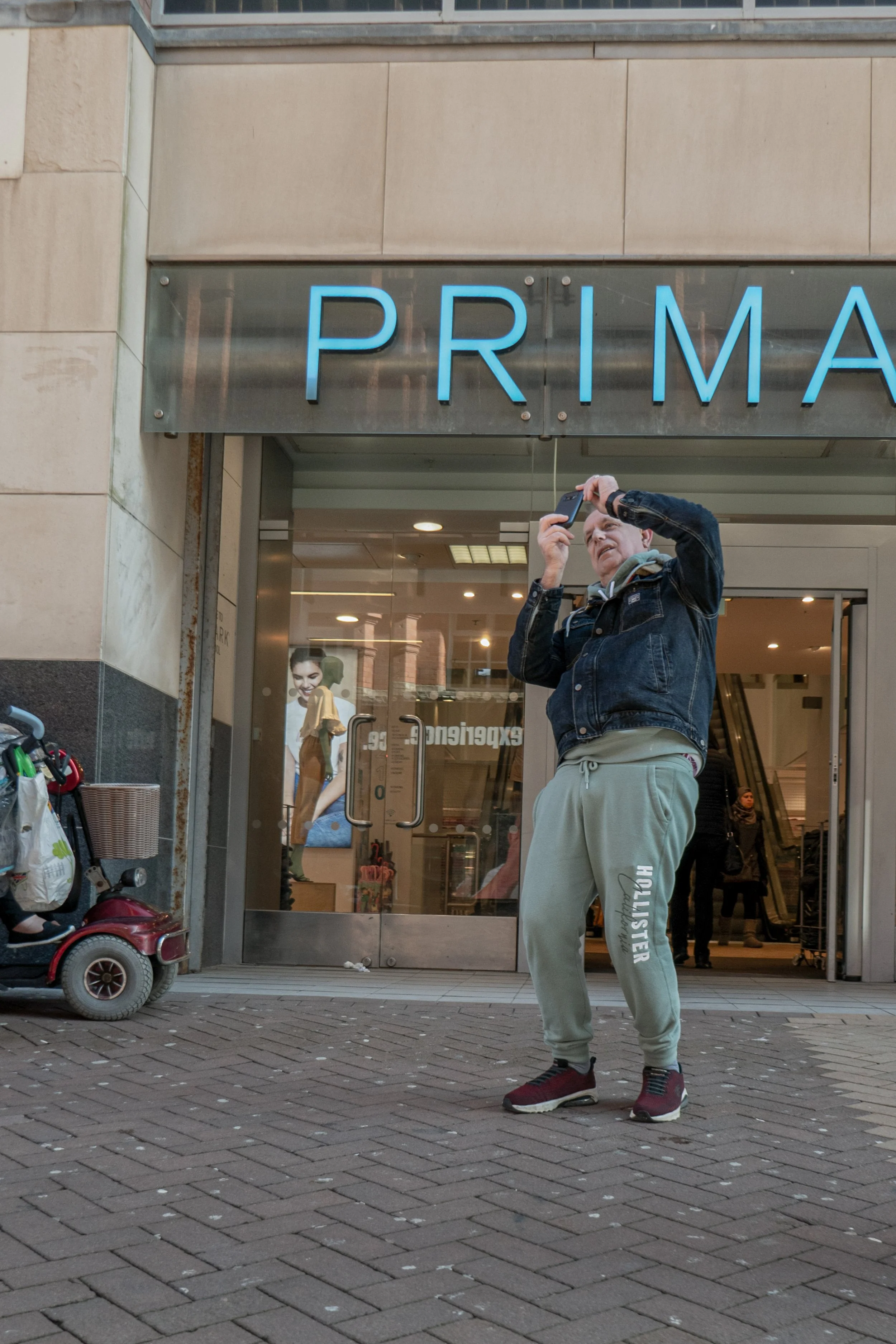 A man taking a selfie in front of a store with a sign that reads 'PRIMA.' He is wearing a black jacket, beige sweatpants with text and graphic prints, and red sneakers. There are shopping carts and a small red wagon on the sidewalk, with a person ins
