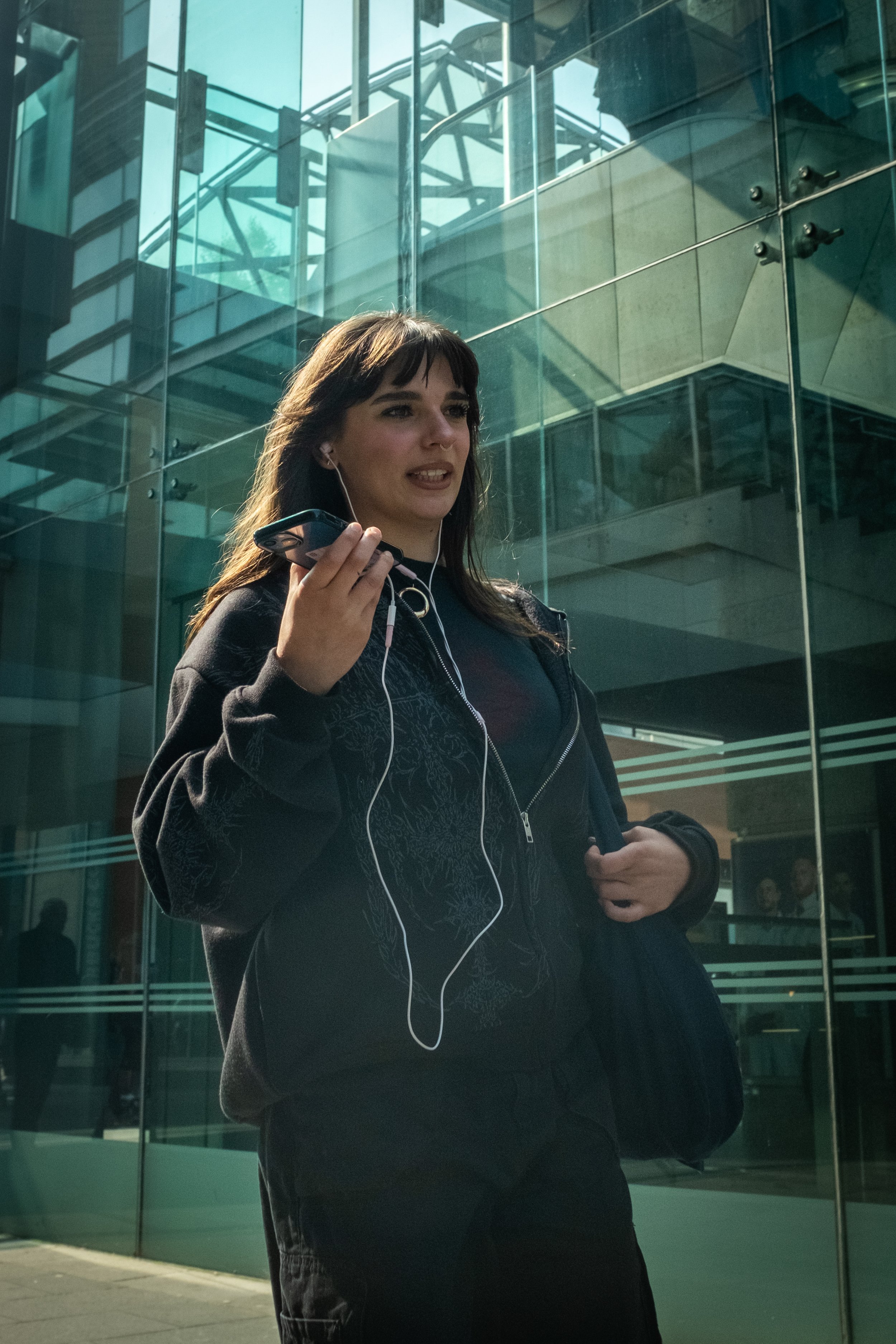 A woman with dark hair and a nose ring is standing outside a modern glass building, holding a smartphone and listening to earphones.