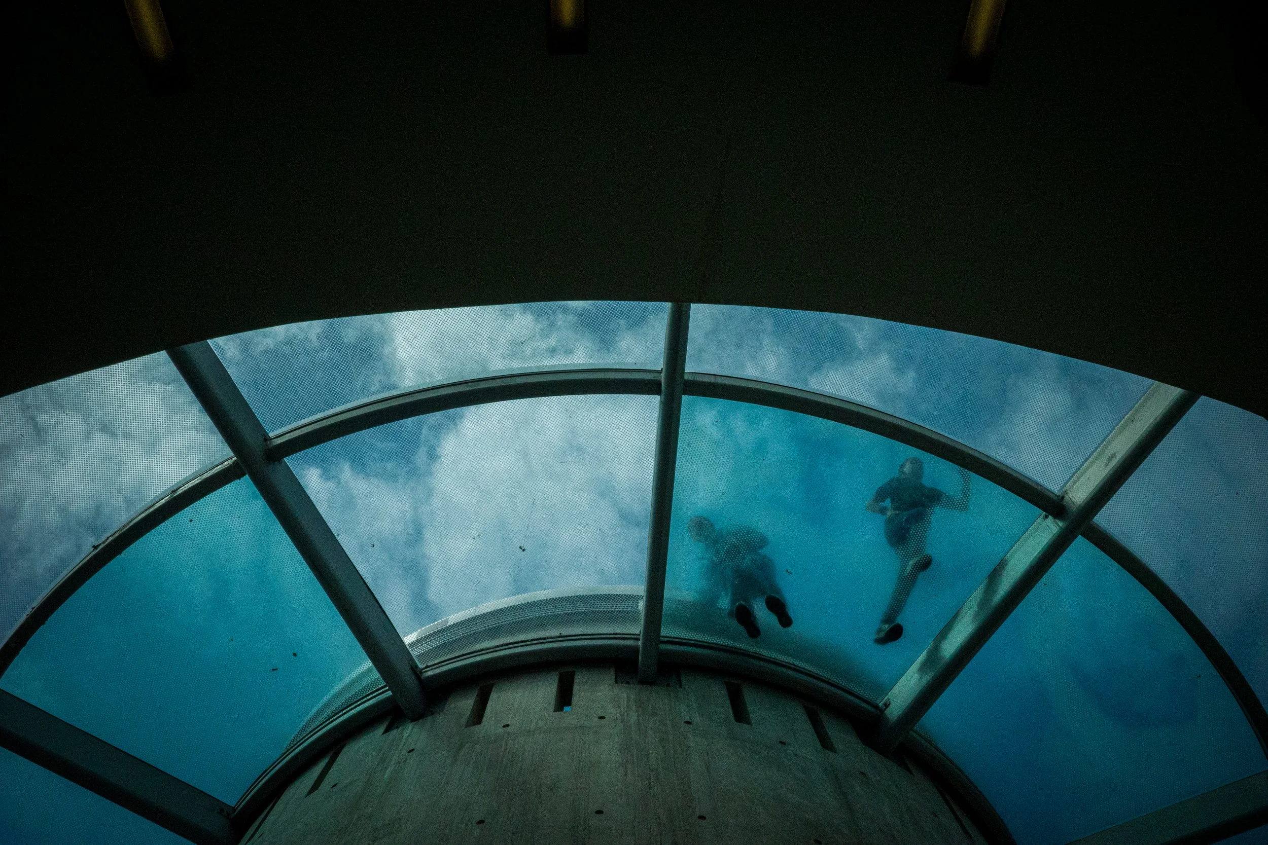 Two people walking on a transparent glass walkway high above the ground, viewed from below with a curved ceiling and cloudy sky in the background.