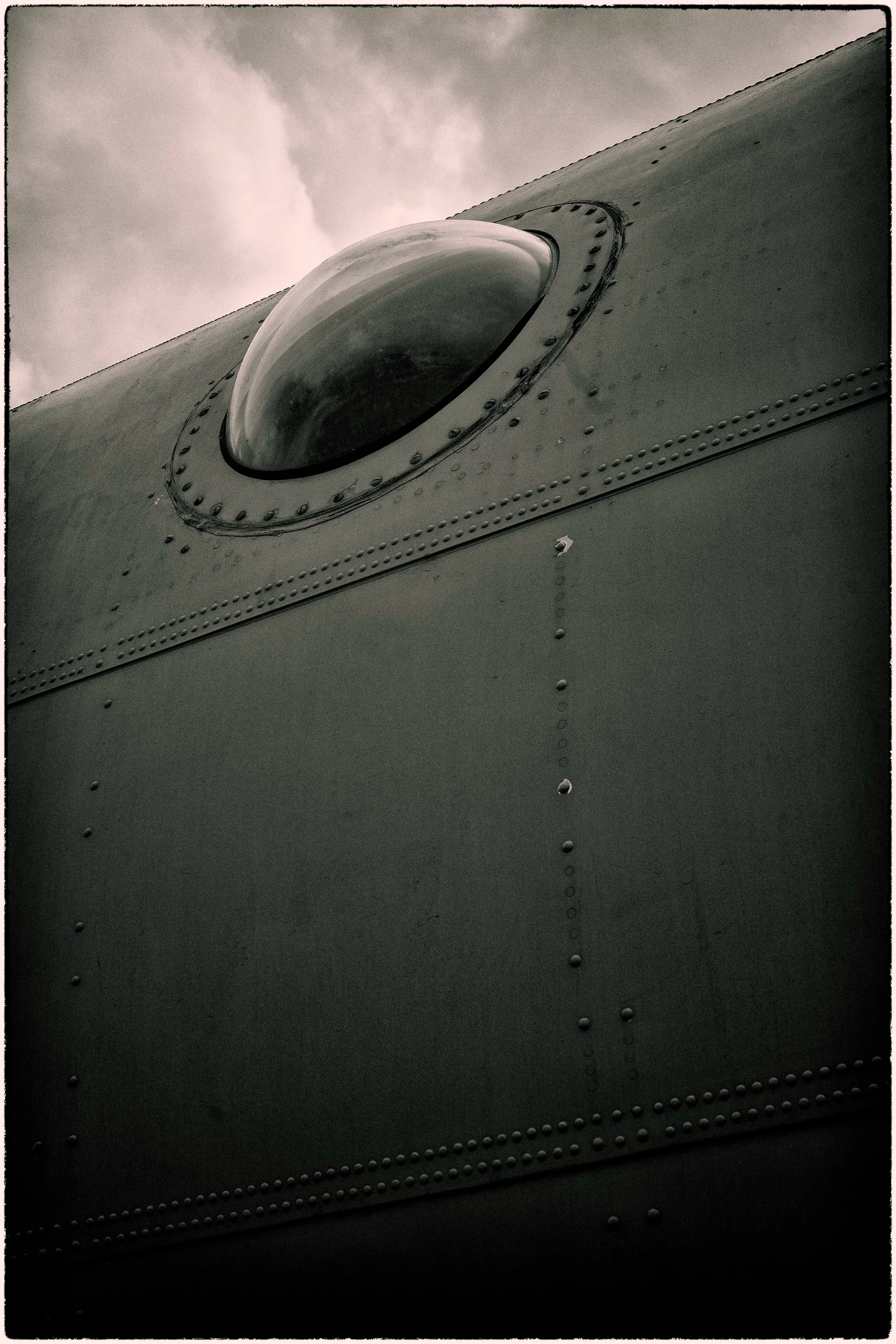 Close-up of an aircraft window or dome, with metal rivets and an overcast sky in the background.
