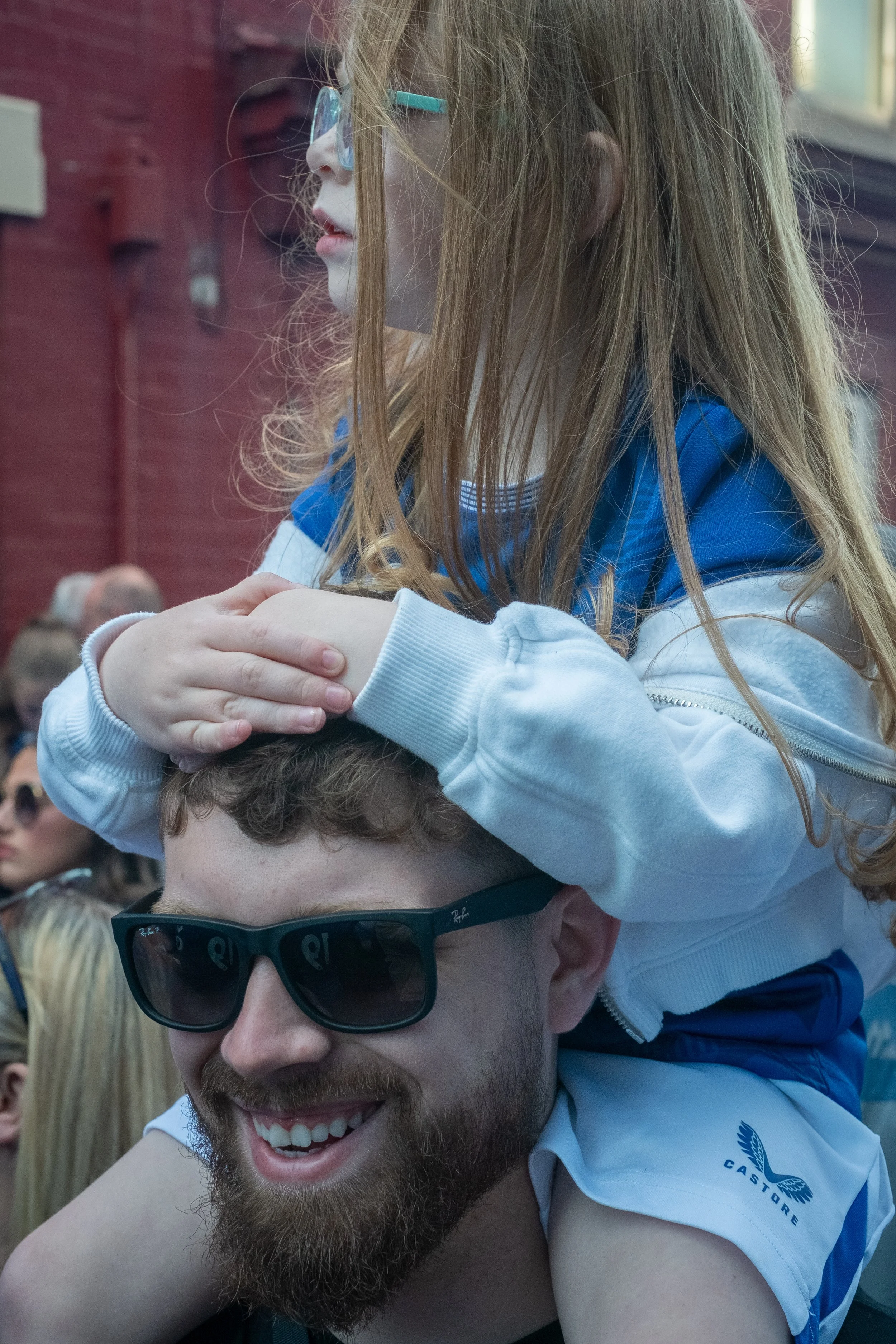A man with a beard and sunglasses smiling, carrying a young girl on his shoulders. The girl has red hair, wears glasses, and is resting her hands on the man's head.