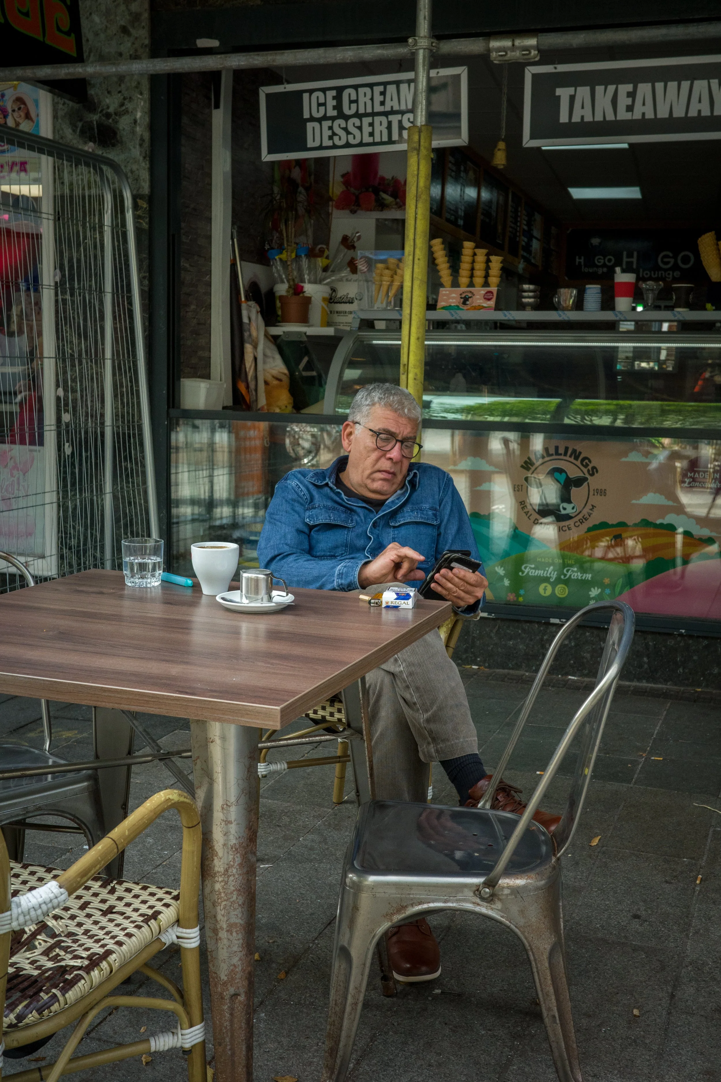 A man with gray hair wearing glasses and a denim jacket sitting at an outdoor café table, looking at his phone. On the table are a glass of water, a cup of coffee, a small metal pitcher, a pack of cigarettes, and a lighter. In the background, there i