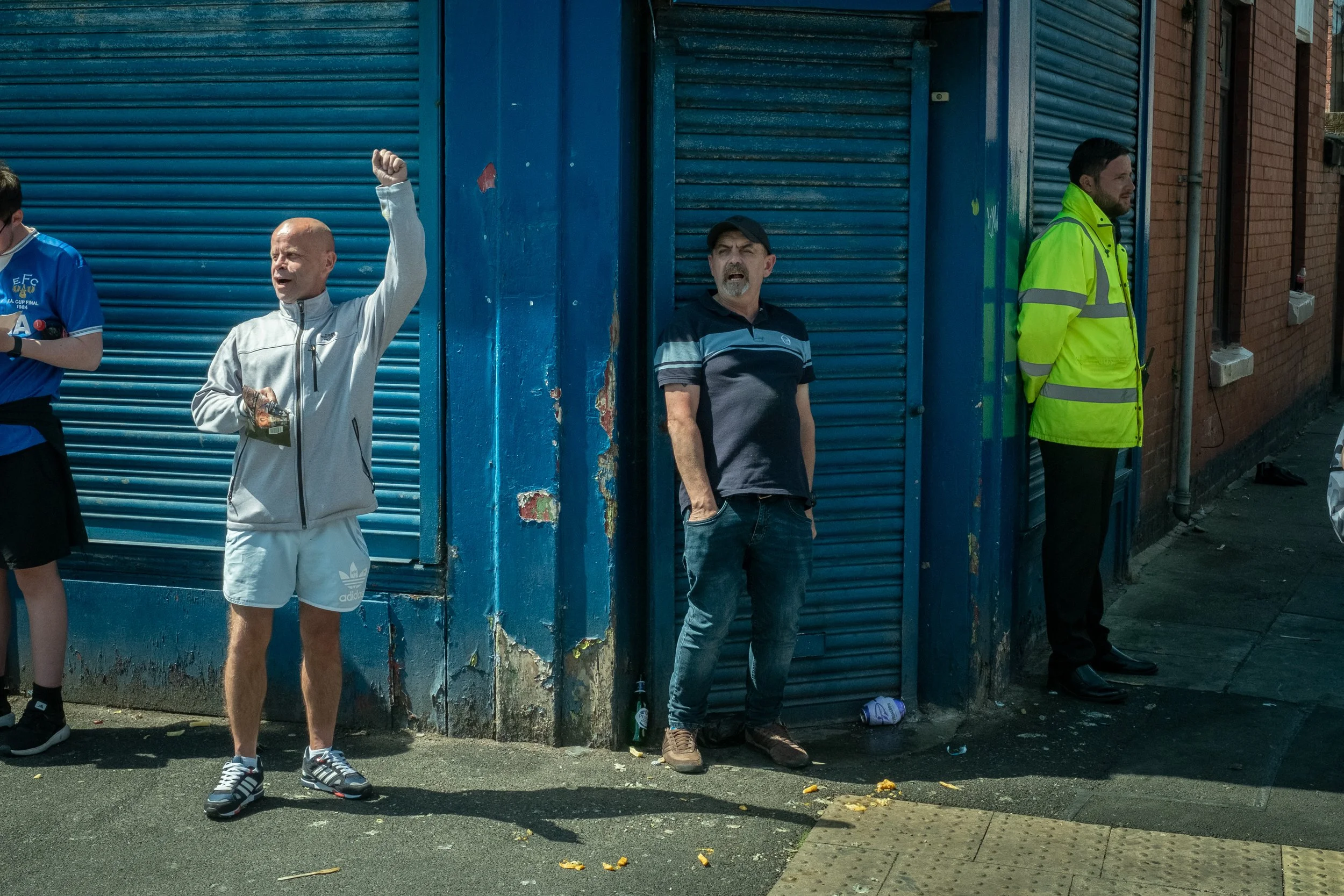 Three men standing on the sidewalk, with blue metal shutters behind them. The man in the middle is wearing a white track jacket and shorts, raising his right arm. The man to his right, leaning against the shutter, is wearing a black and gray polo shi
