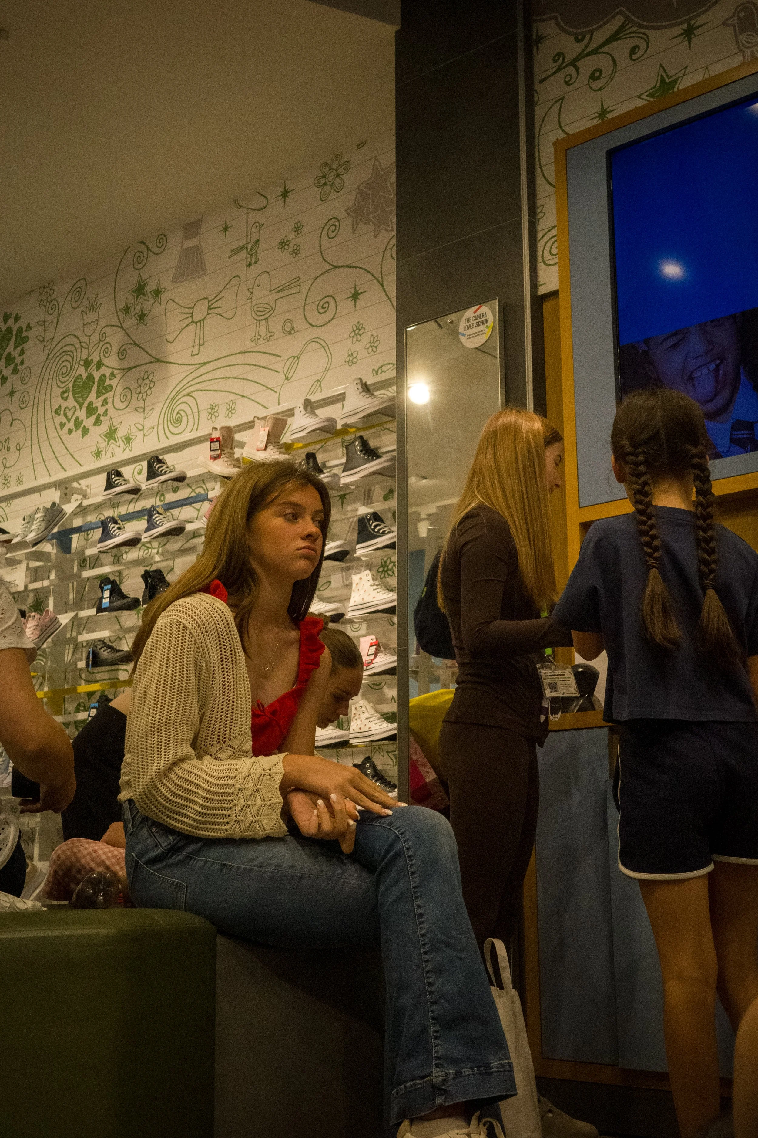 Young women and girls standing and sitting in line at a store checkout area with colorful wall art and shoe displays in the background.