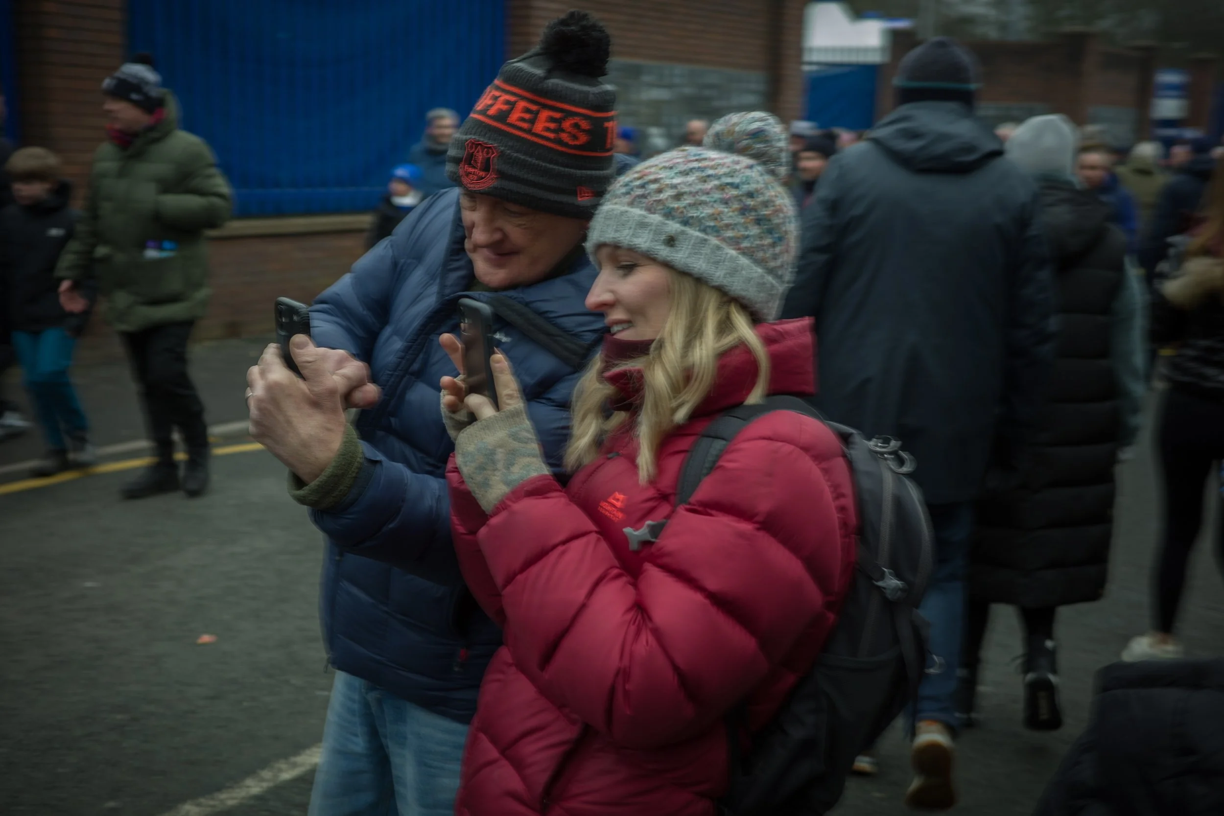 Two people wearing winter clothing, including hats, are standing close together and looking at a smartphone during a gathering outdoors with other people in the background.