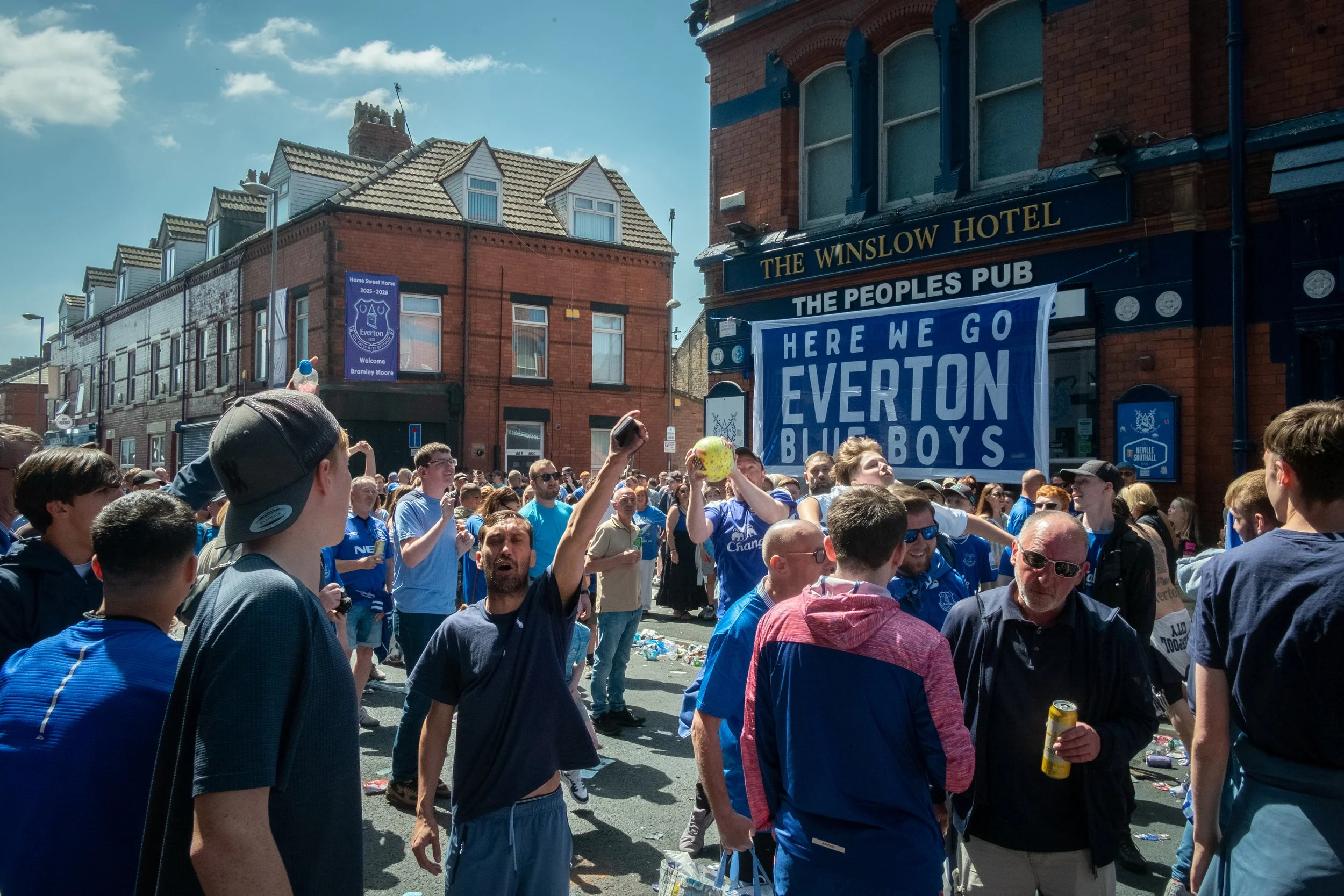 Crowd of Everton Football Club supporters gathered outside a pub with a large blue banner that reads 'Here We Go Everton Blue Boys.' Many are wearing blue jerseys, some are taking pictures or holding drinks, and the sky is clear and sunny.