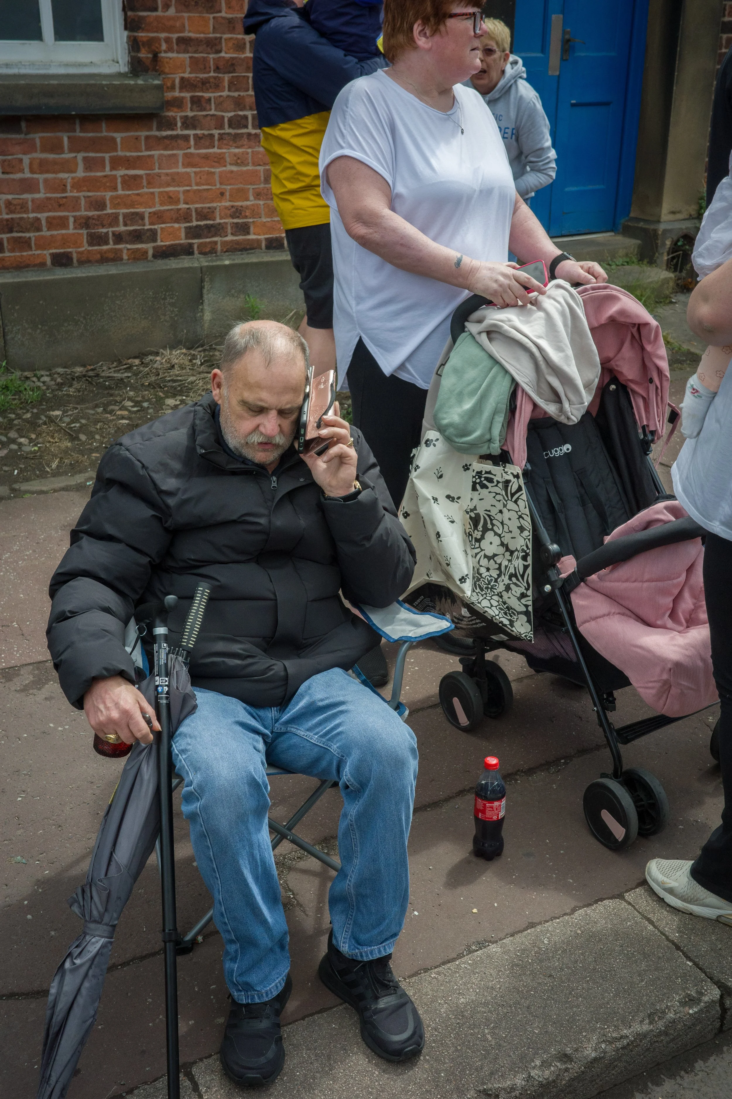 A man with glasses and a gray beard sitting in a wheelchair, talking on a cellphone, with a bottle of Coca-Cola on the ground in front. Behind him, a woman with short red hair and glasses pushes a stroller, and several other people are standing nearb
