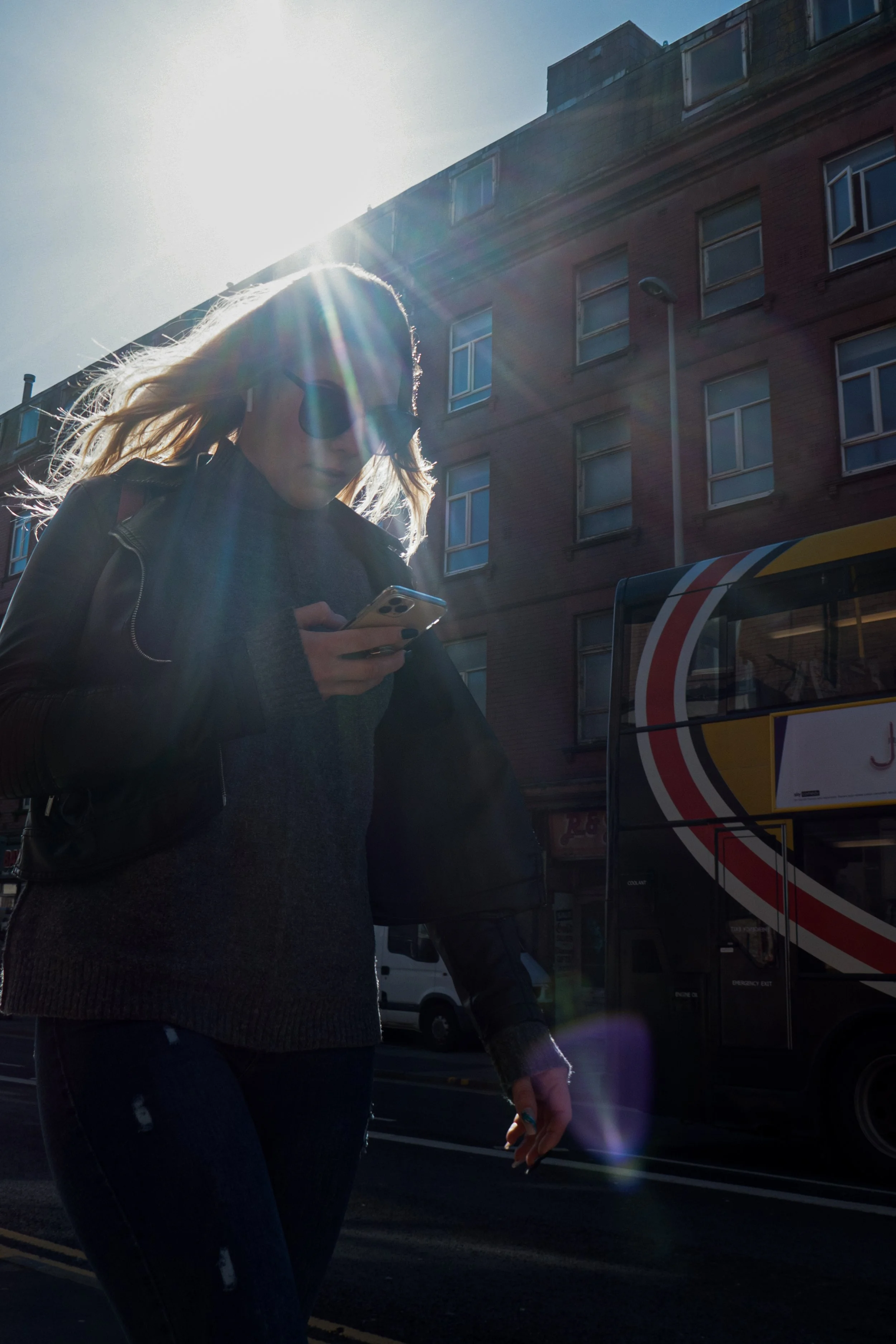 A woman wearing dark sunglasses, a black leather jacket, and distressed jeans is walking outside on a sunny day, looking at her phone. The sun is behind her, creating lens flare and casting her shadow on the street. Behind her is a red brick building