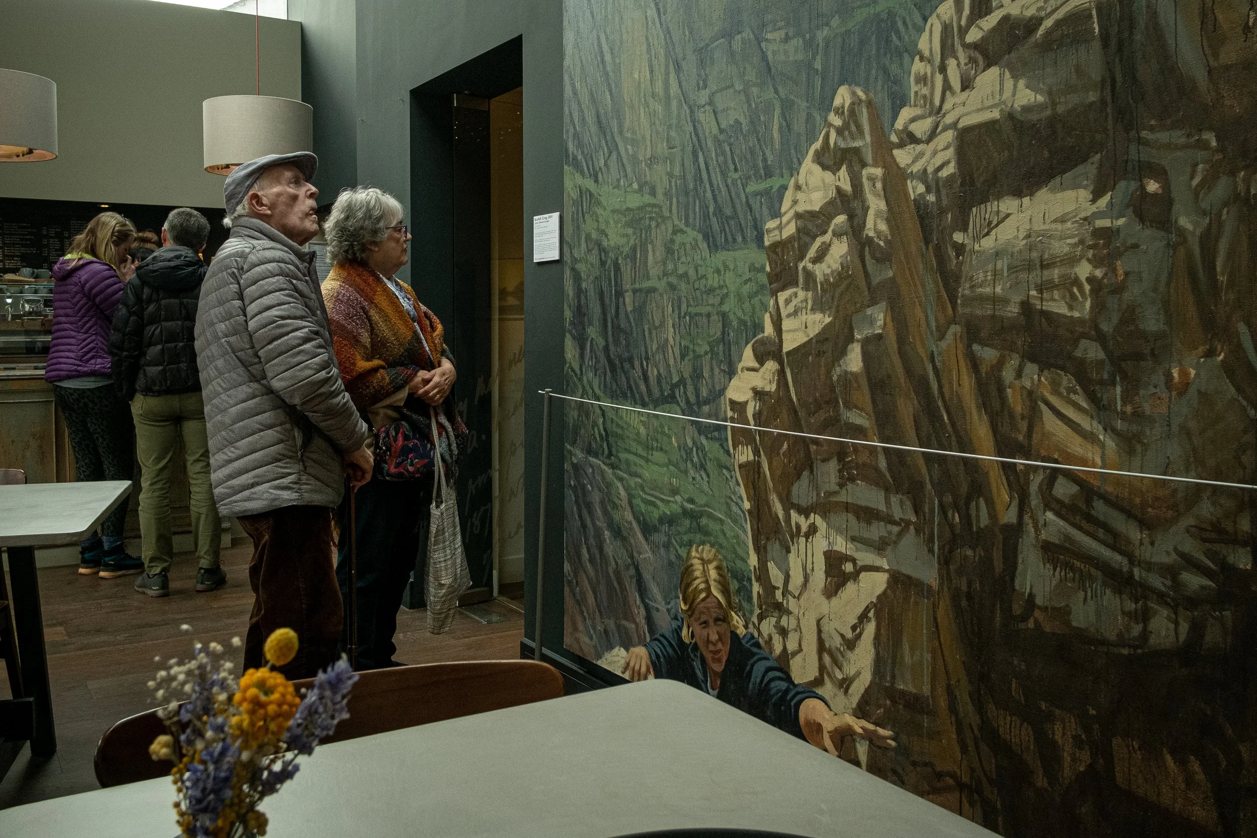 Two people observing a large mural of a mountainous landscape inside a cafe, with other patrons in the background and a small bouquet of flowers on a table in the foreground.