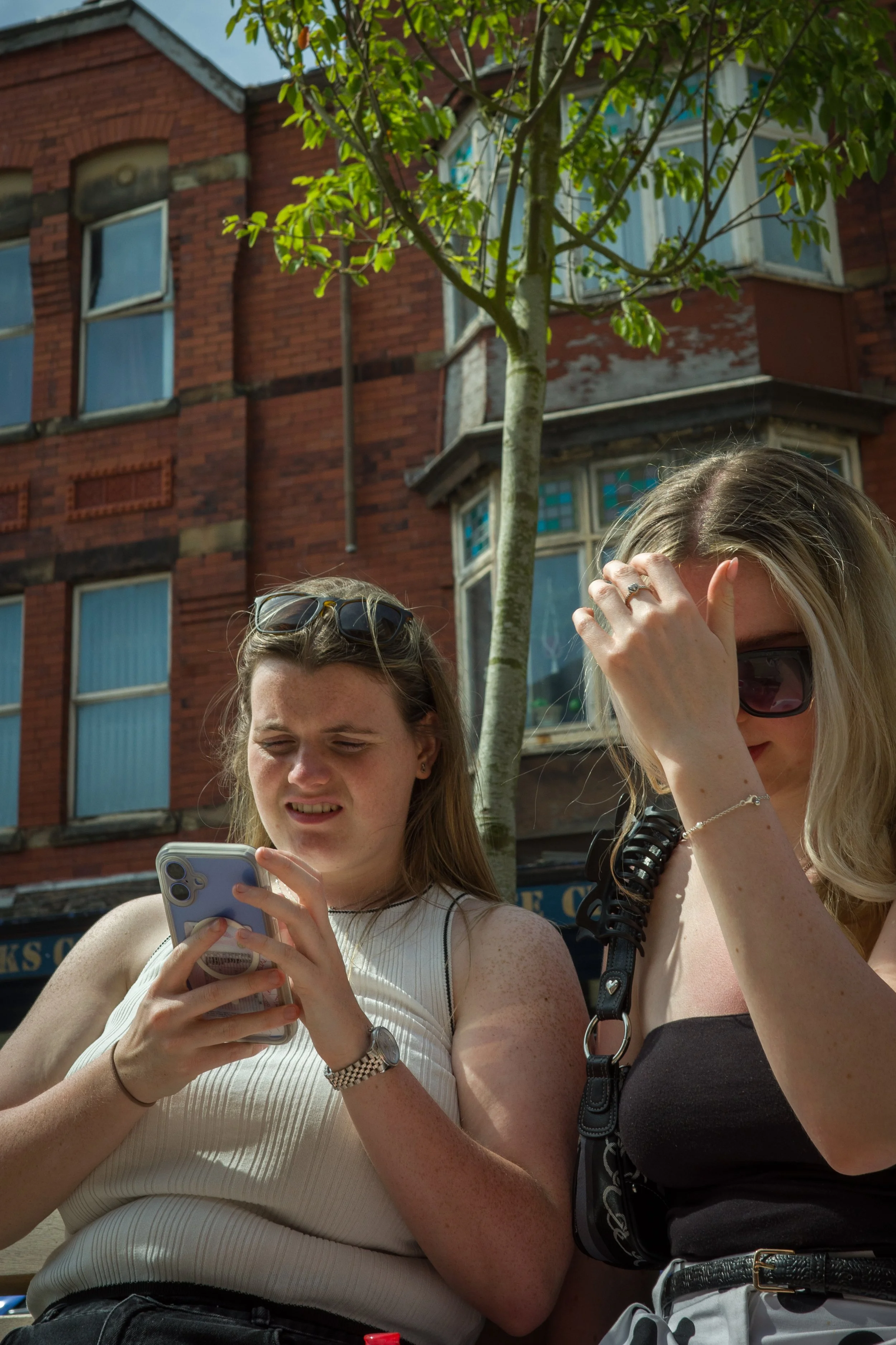 Two young women standing outdoors in front of a red brick building with trees, one is looking at her phone, and the other is adjusting her sunglasses.