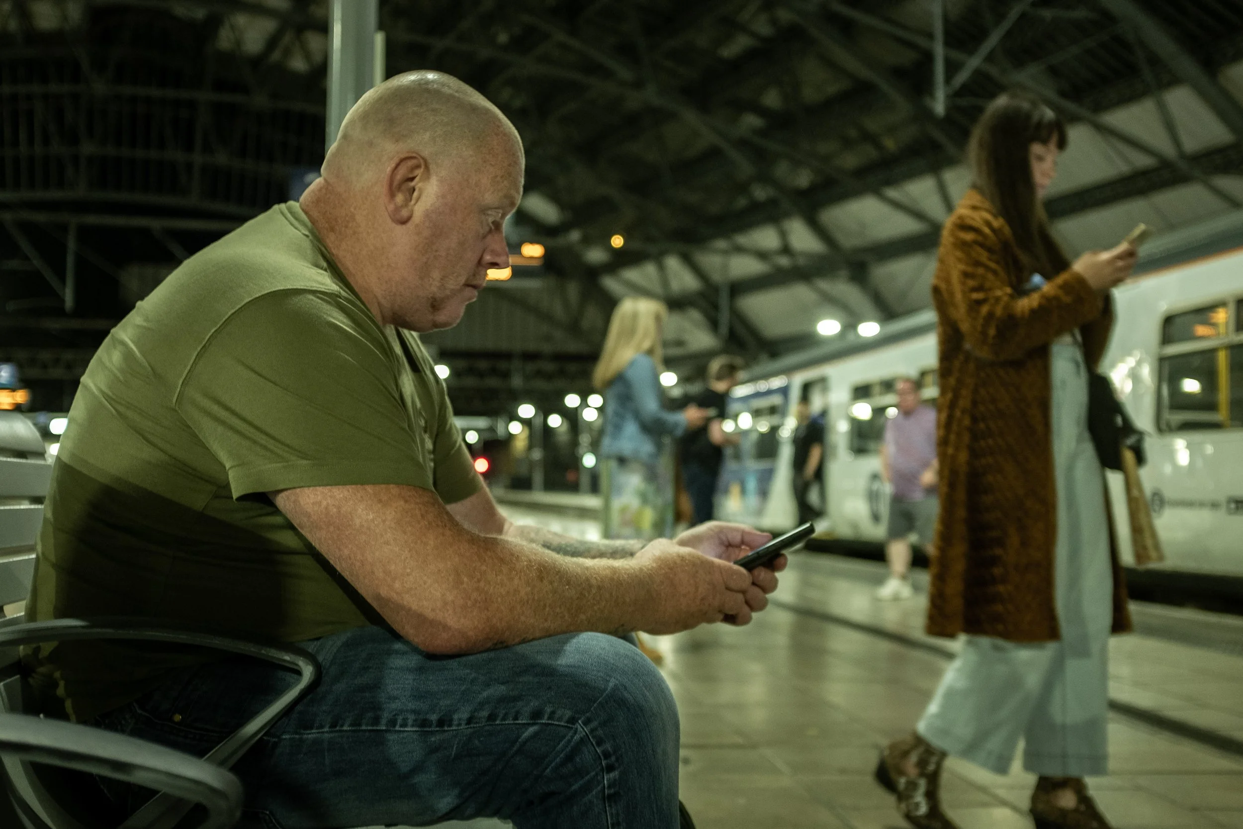 A man with a shaved head wearing a green t-shirt sitting on a bench at a train station, looking at his phone. Several other people are in the background, some standing and some waiting for the train, with a train visible at the platform.