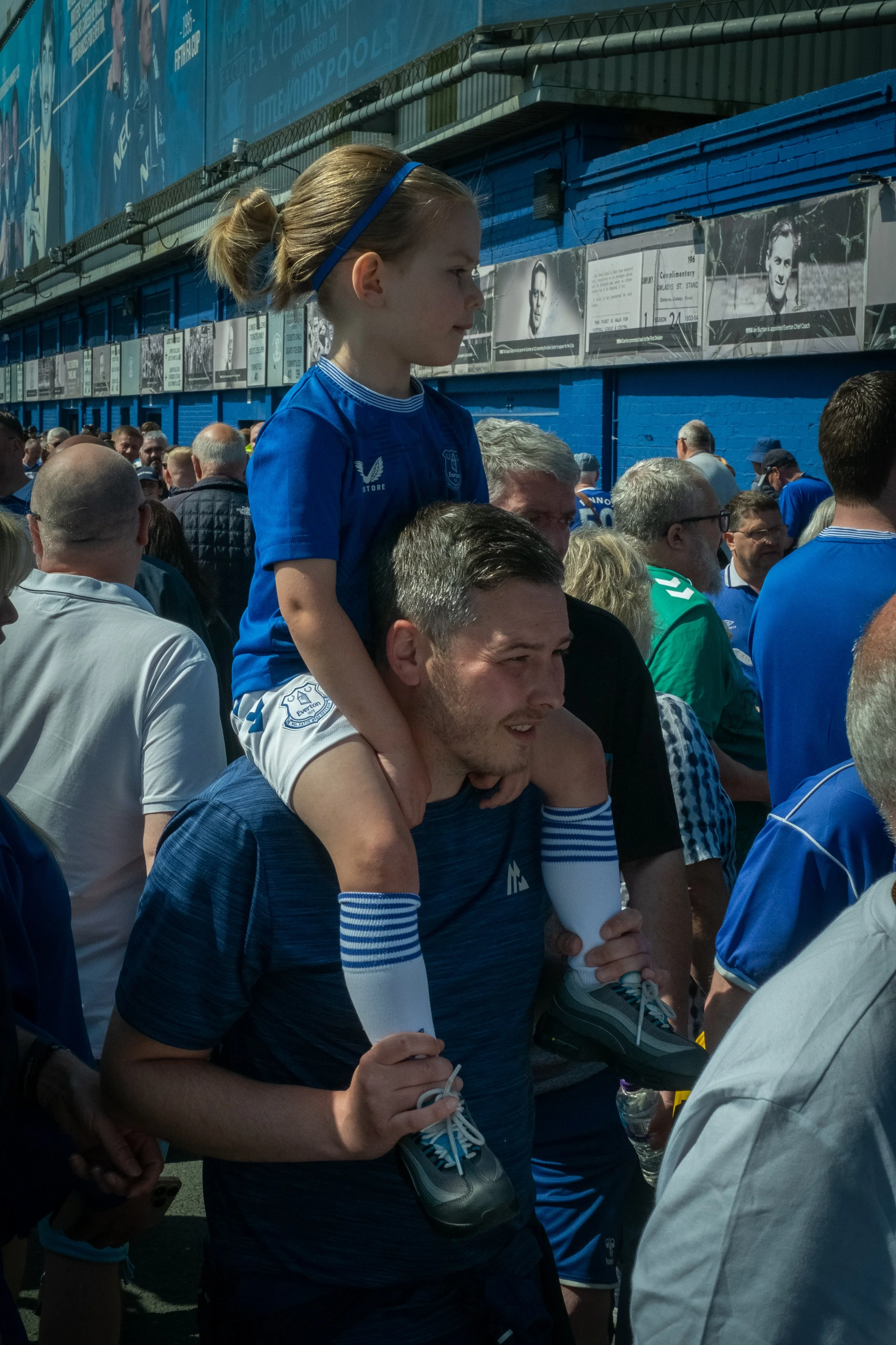 A man carrying a young girl on his shoulders in a crowded outdoor area with blue painted walls and banners with images of women and text.