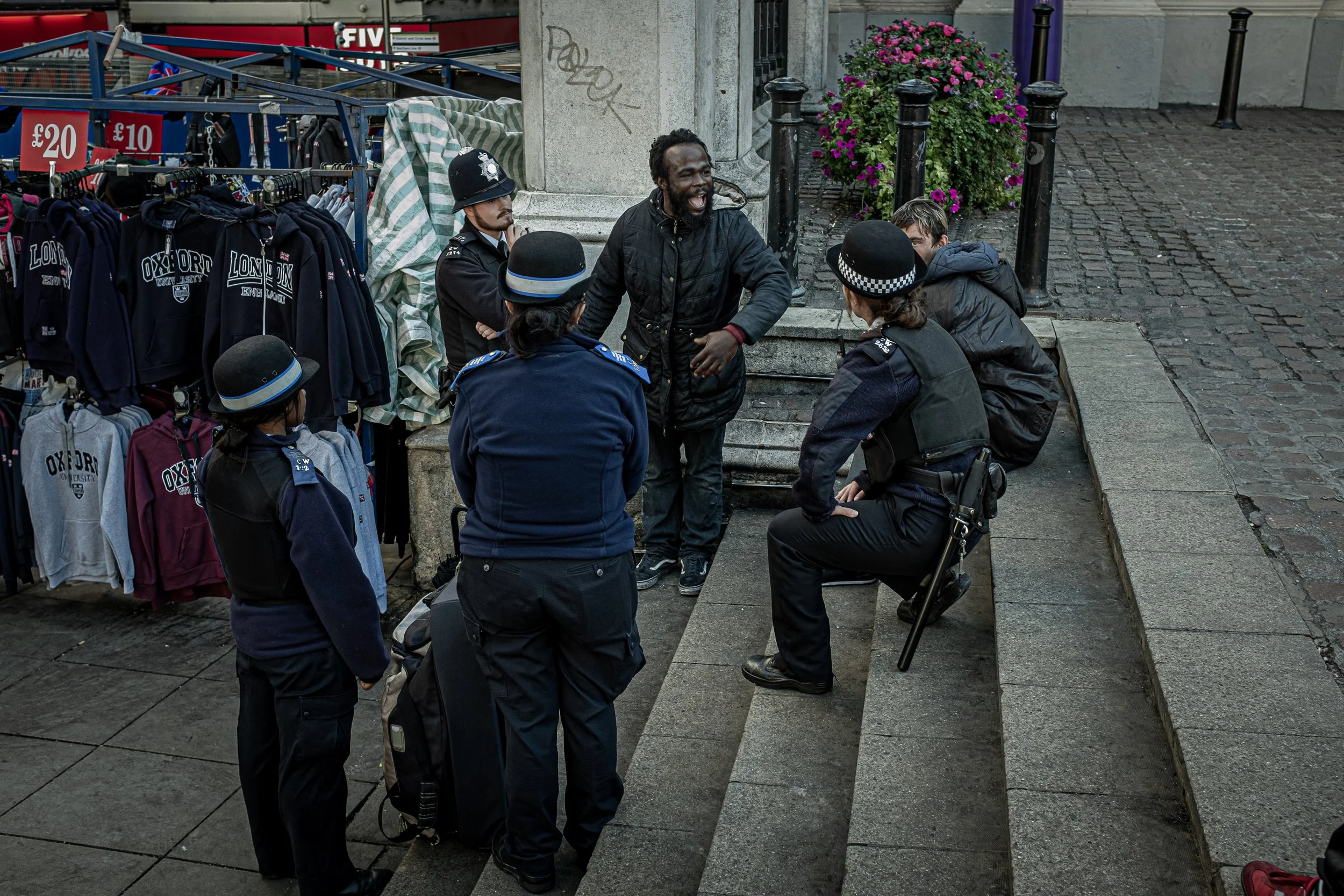 A man with dreadlocks and a beard is talking with a group of police officers at a street corner near a clothing stall. The police officers are in uniform, some wearing hats, one sitting on steps, and the others standing. The street has cobblestones, 