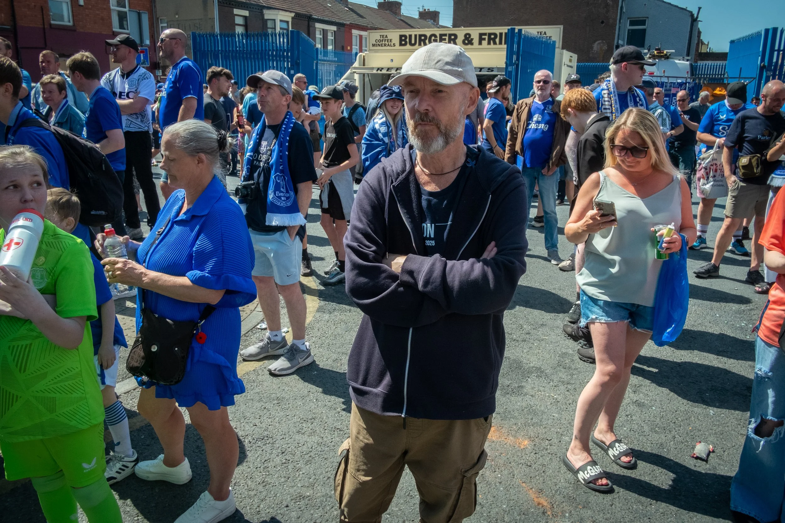 Crowd of football fans gathered outside, many wearing blue jerseys, scarves, and hats, with some holding drinks and phones, near a blue fence and food stand.