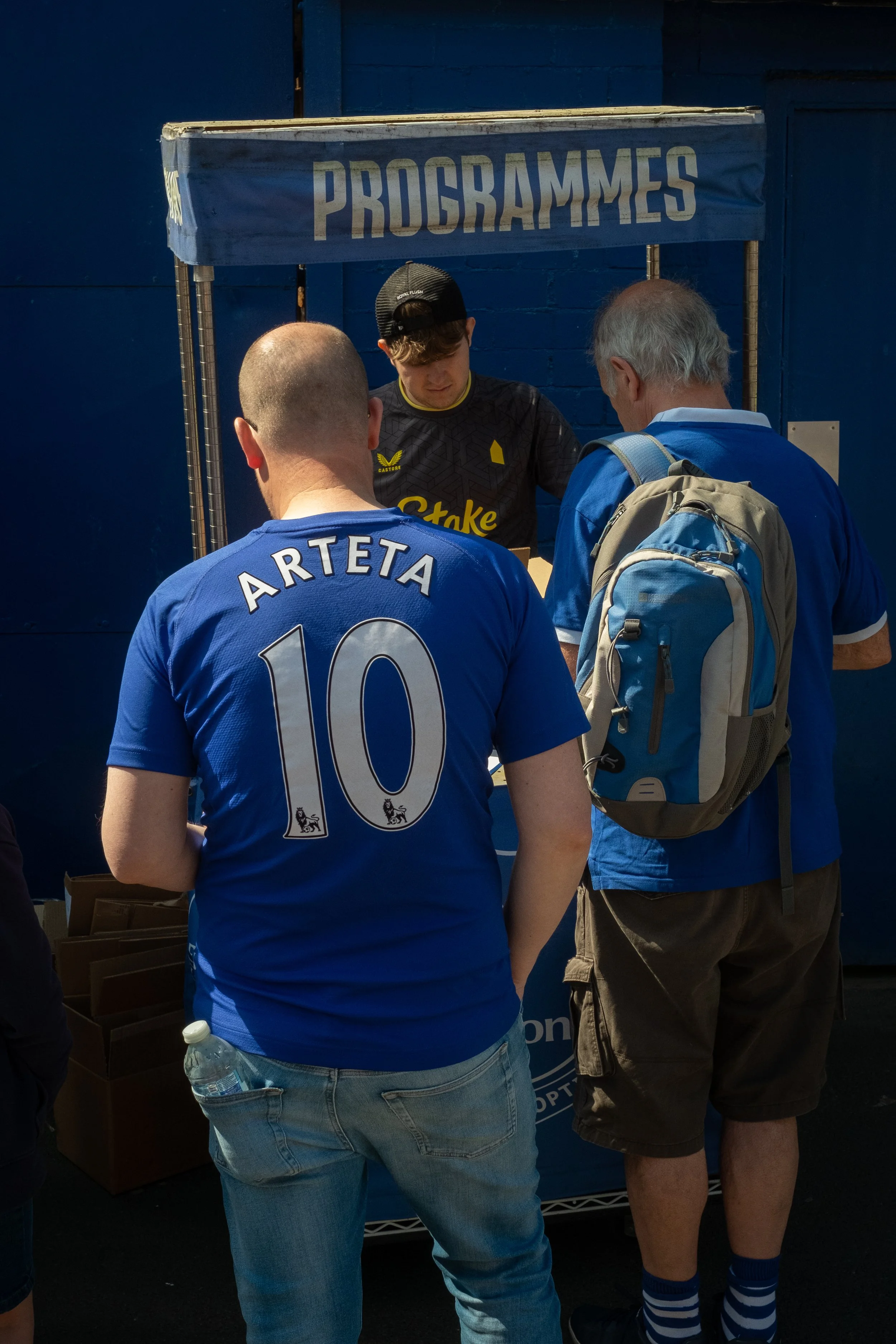 Men standing at a program booth with a sign above, one wearing a blue soccer jersey with number 10 named ARTETA, another with a blue backpack, and a young man behind the booth.