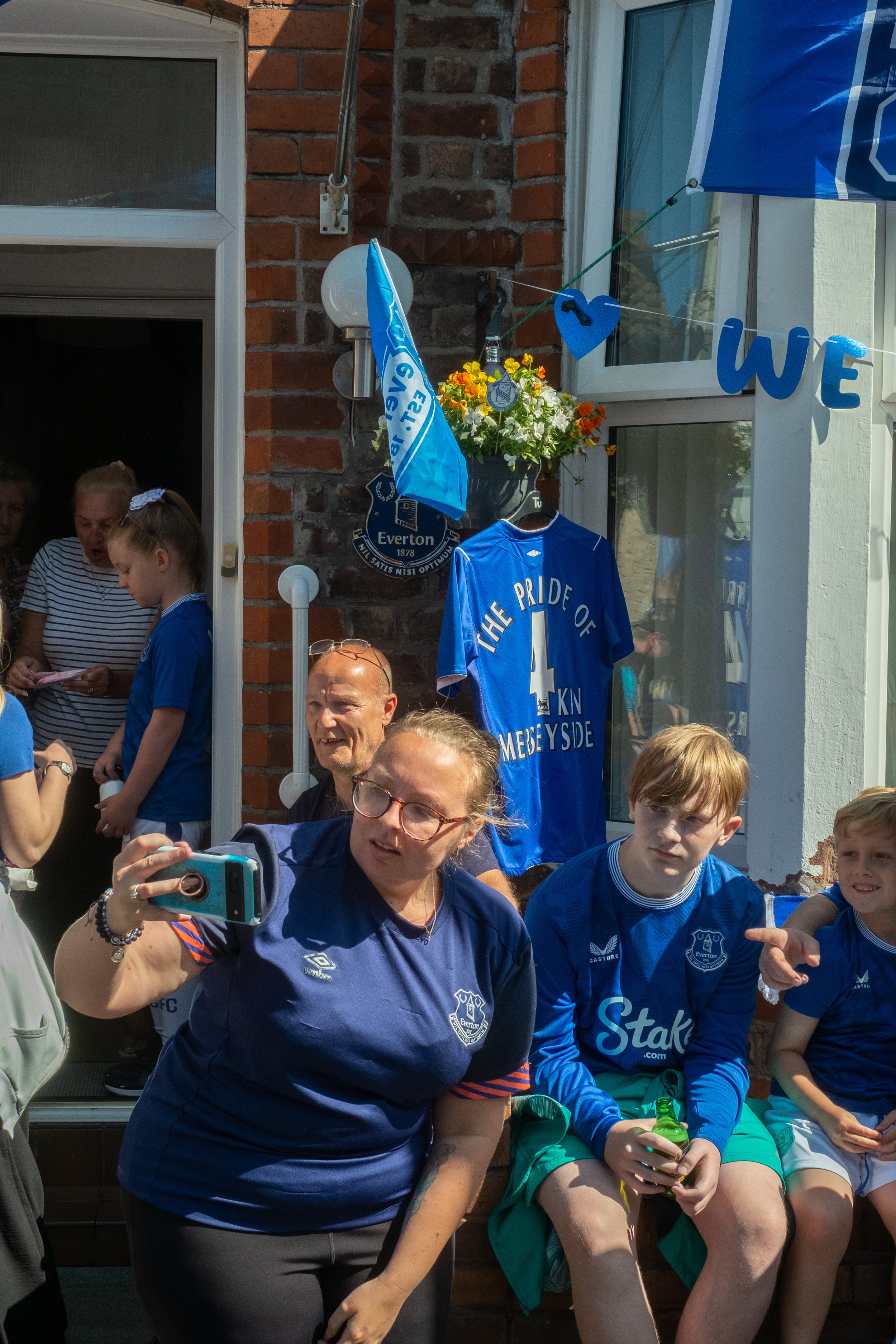 Group of people, including children and adults, celebrating outside a house decorated with Everton Football Club banners and jerseys, with a prominent jersey displaying 'The Pride of Merseyside' and the number 4