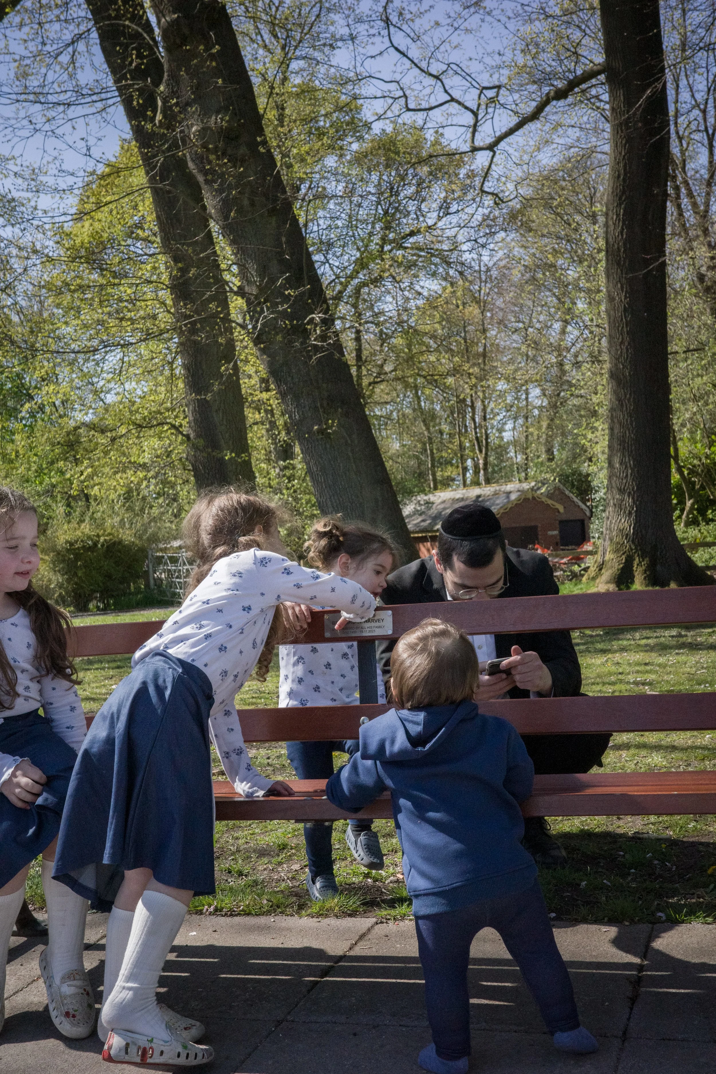 A group of children and an adult gathered around a bench outdoors, with trees and a small building in the background, engaging with a smartphone.