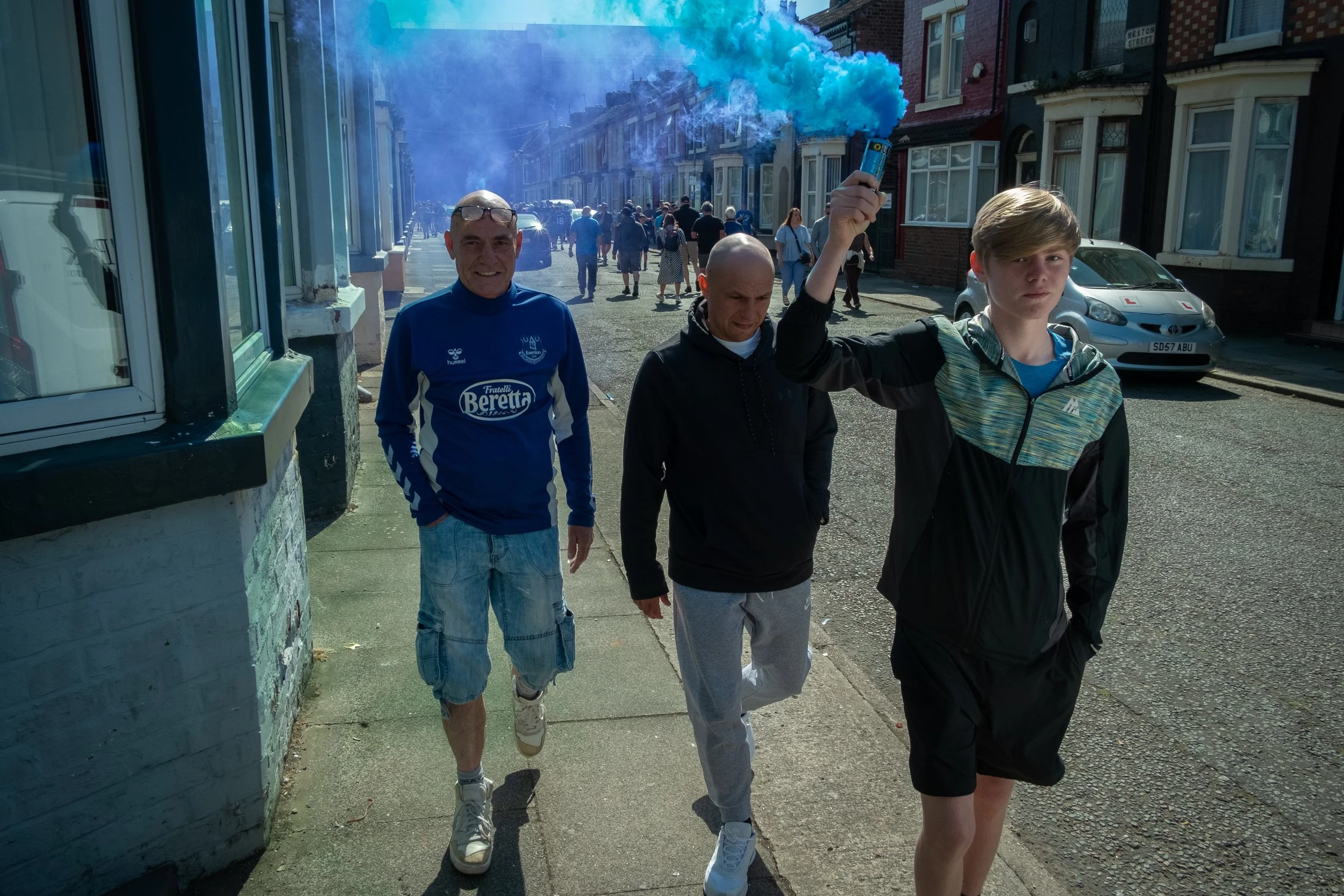 Three young men walking along a street, one holding a blue smoke flare, with people and buildings in the background.