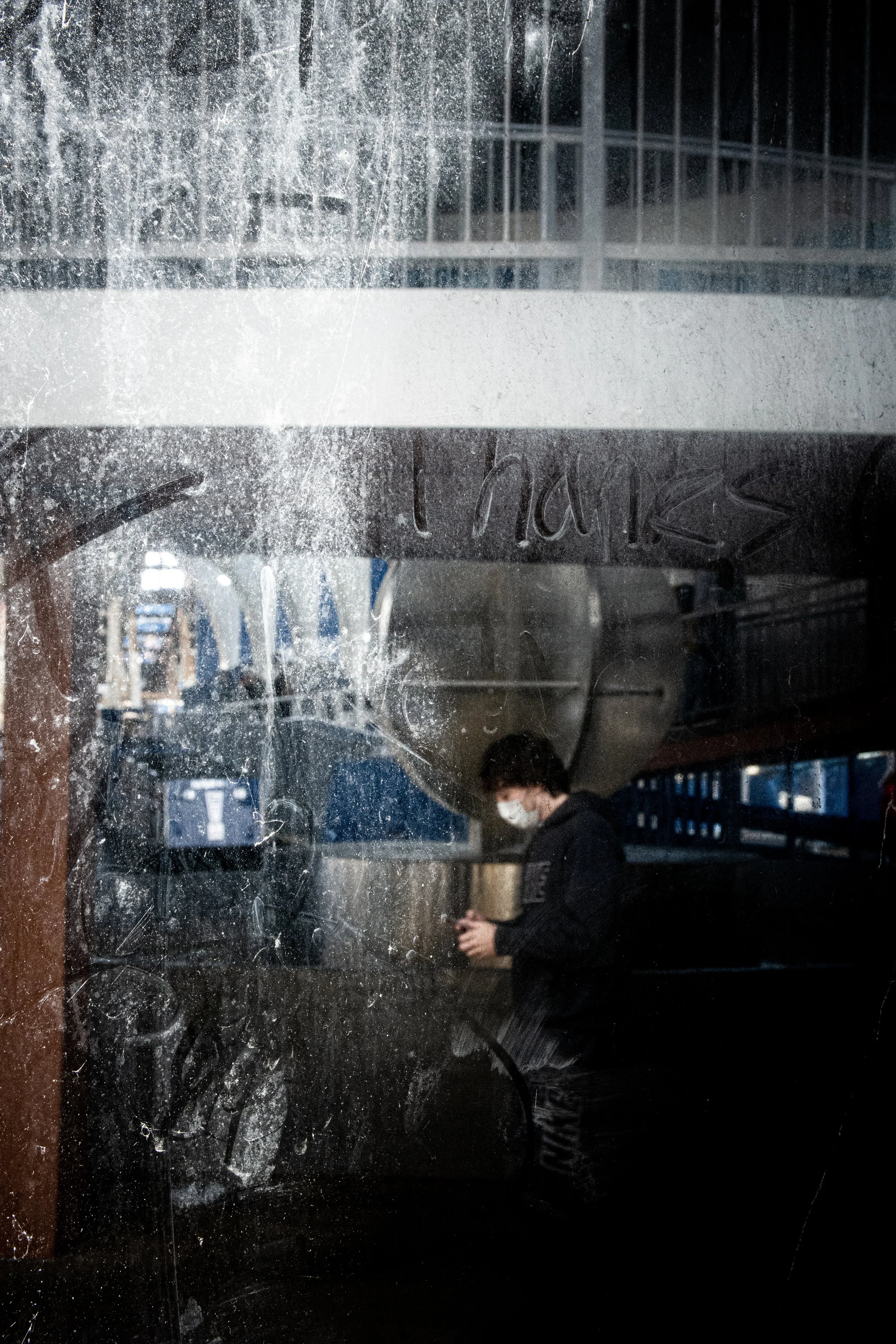 A person wearing a face mask and black clothing standing in front of a dirty glass window with scratches and smudges, holding an object in their hands. The background shows an indoor setting with some metal shelving and bright blue and white decor.