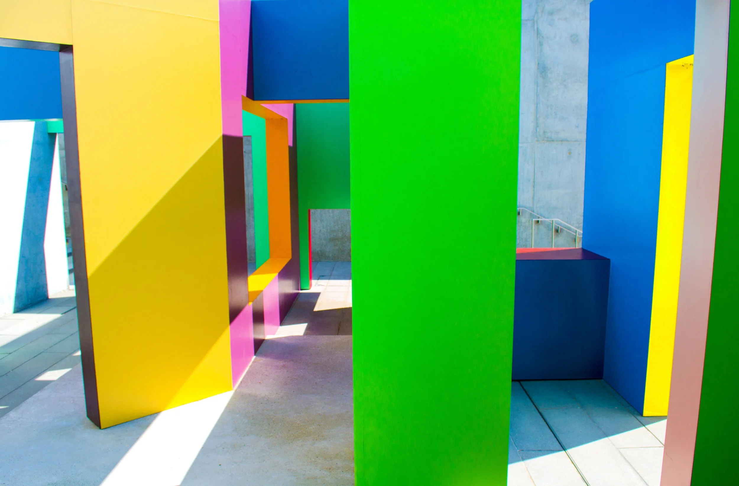 Colorful geometric sculptures with large, flat, painted panels in yellow, pink, blue, green, red, and beige, arranged in a pattern outdoors.