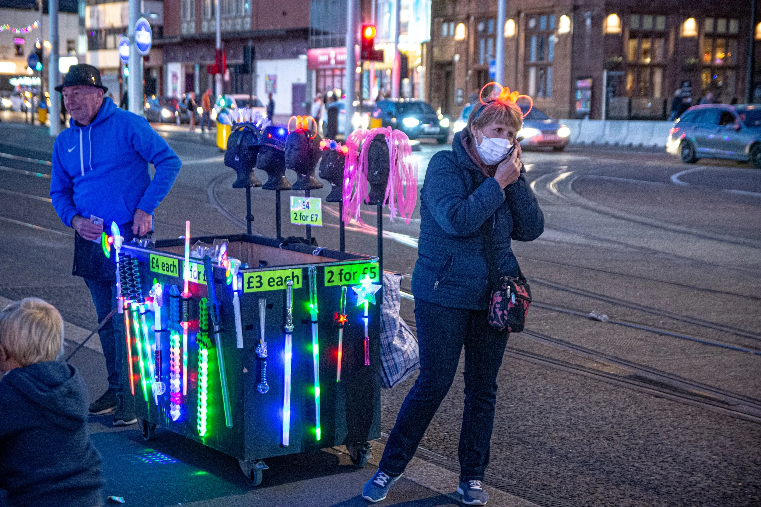 A street scene at dusk with people, a stand selling illuminated glow sticks and accessories with signs indicating prices, and cars driving by. One woman is wearing a face mask and glow stick headband, possibly shopping or browsing.