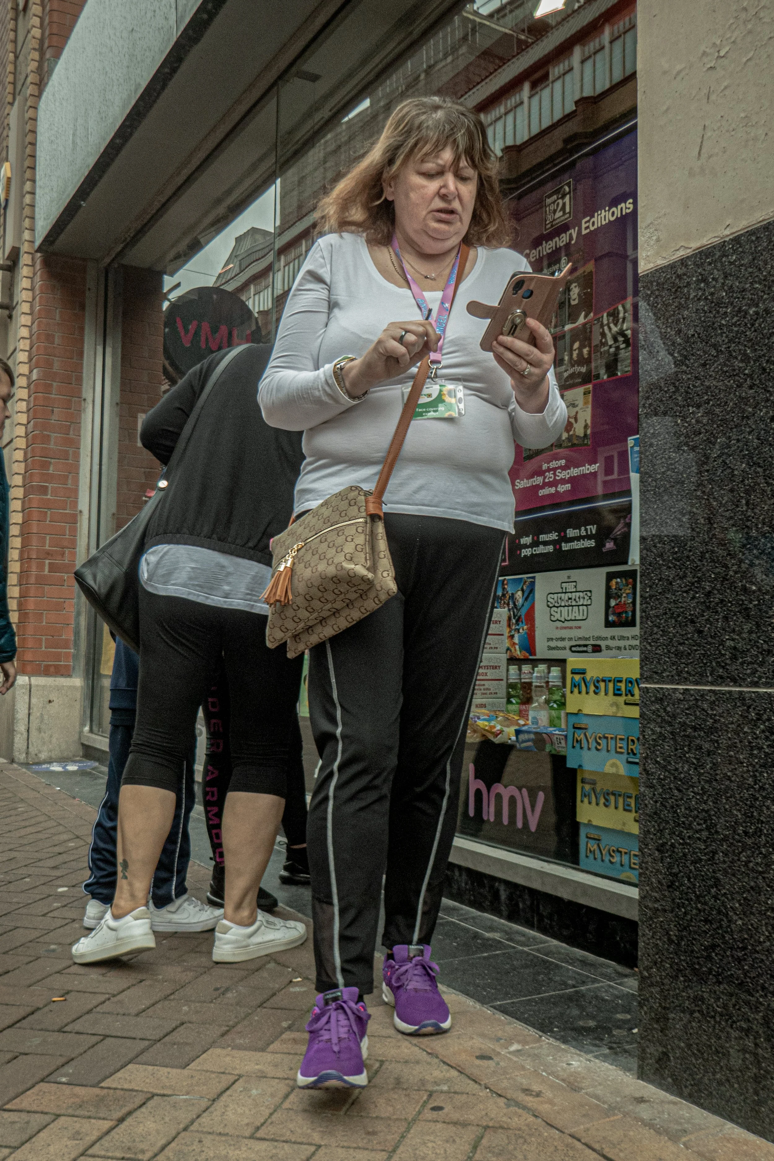 A woman with curly hair wearing a white long sleeve shirt, black pants with white stripes, and purple sneakers, looking at her phone while standing outside a store with other people nearby.