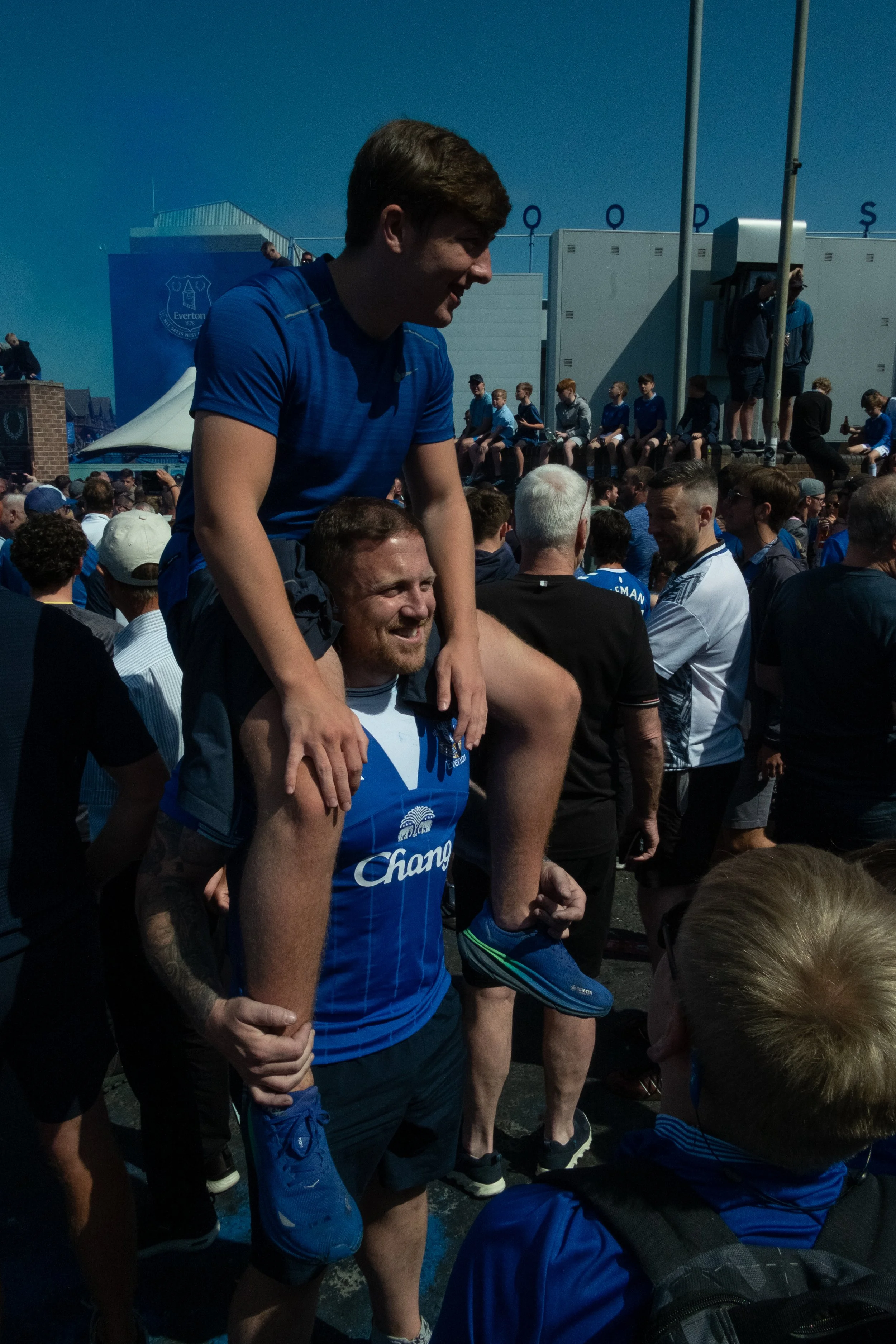 Two men celebrating, one carrying the other on his shoulders at a sports event with a crowd and players in the background