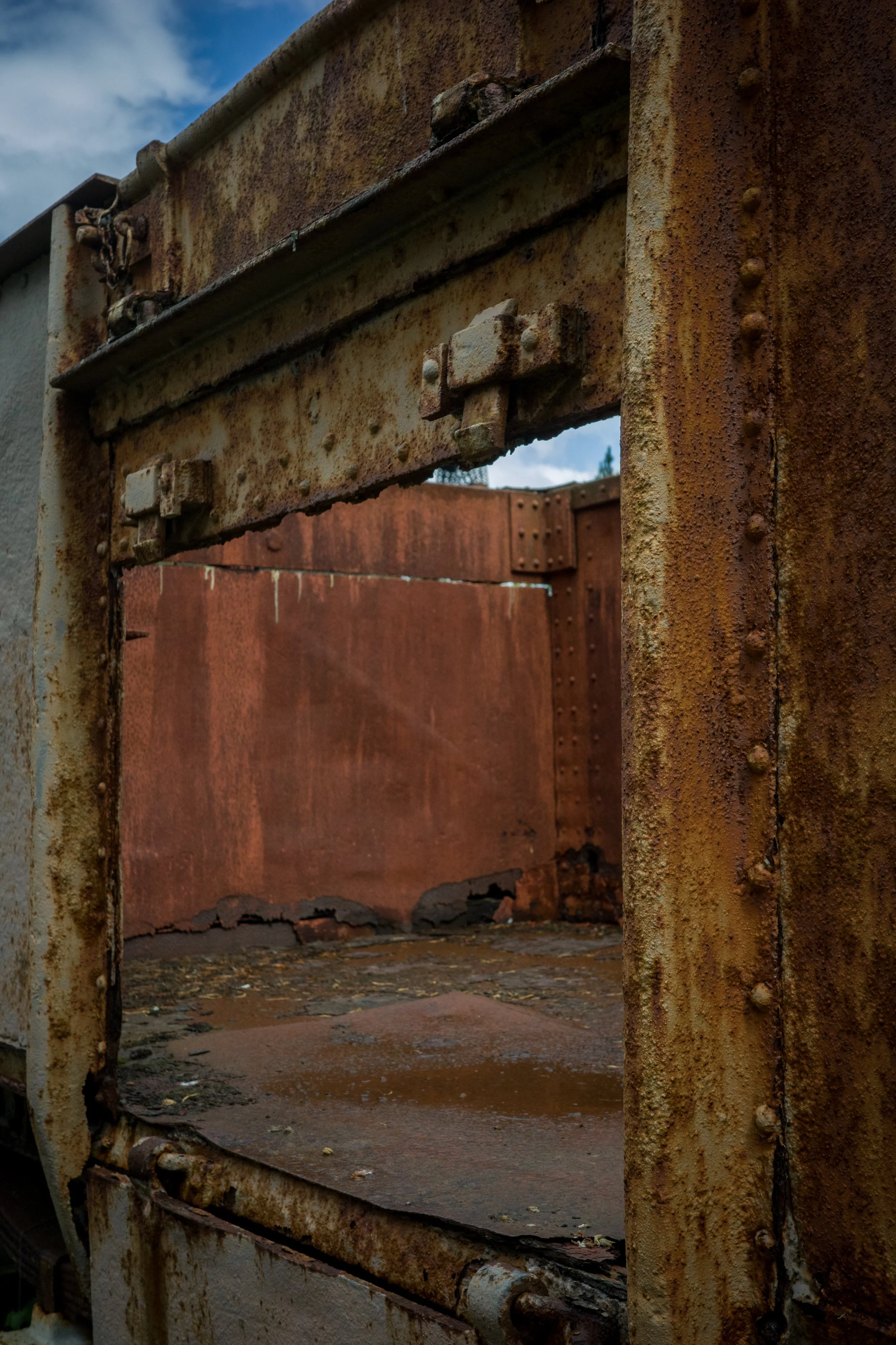 Close-up of a corroded metal gate frame and hinges, showing rust and peeling paint, with a background of a rusted metal sheet and a partly cloudy sky.