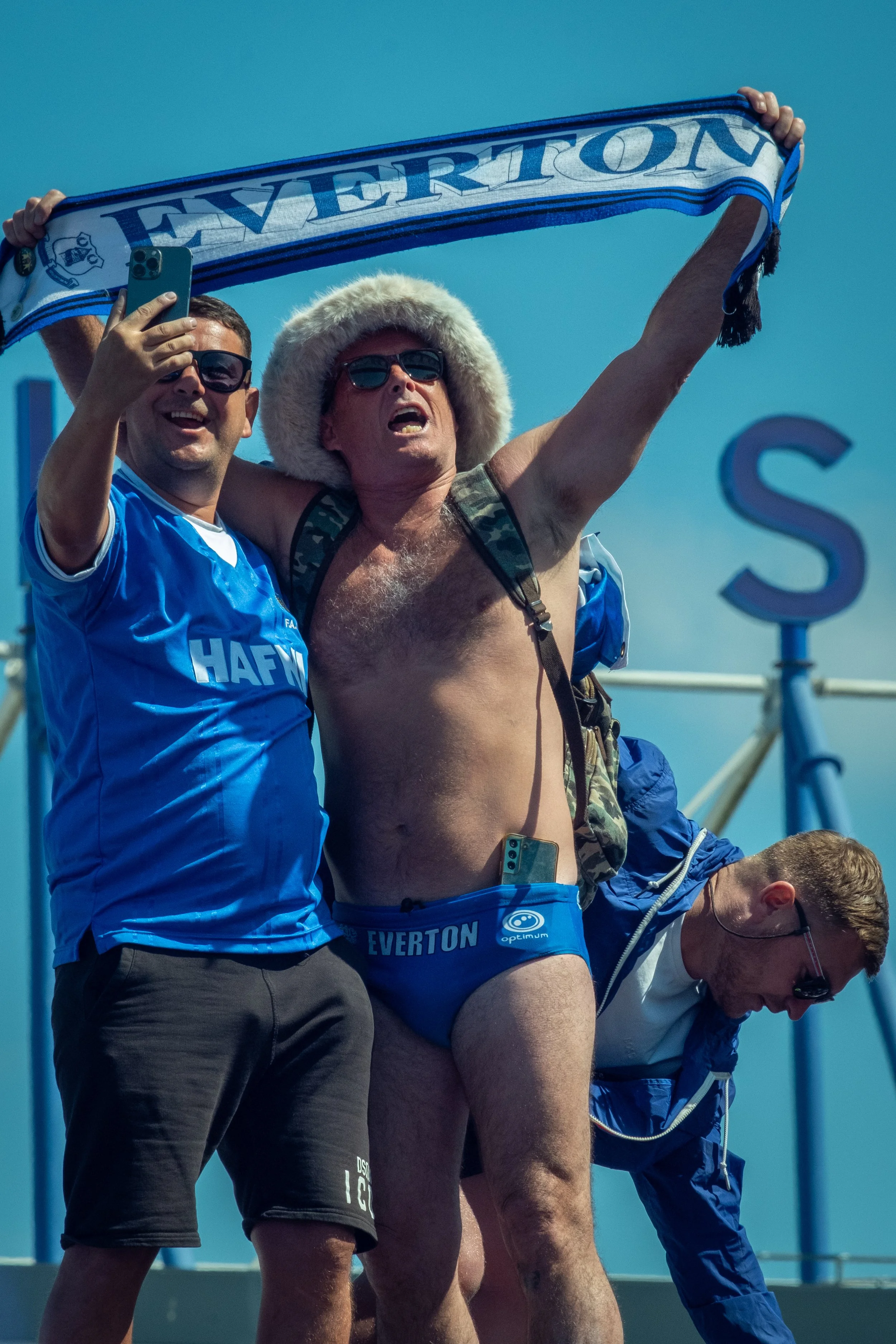 A group of men celebrating, with one man dressed in blue Everton football fan gear, holding a scarf, and wearing only underwear and sunglasses, with others taking selfies and smiling.